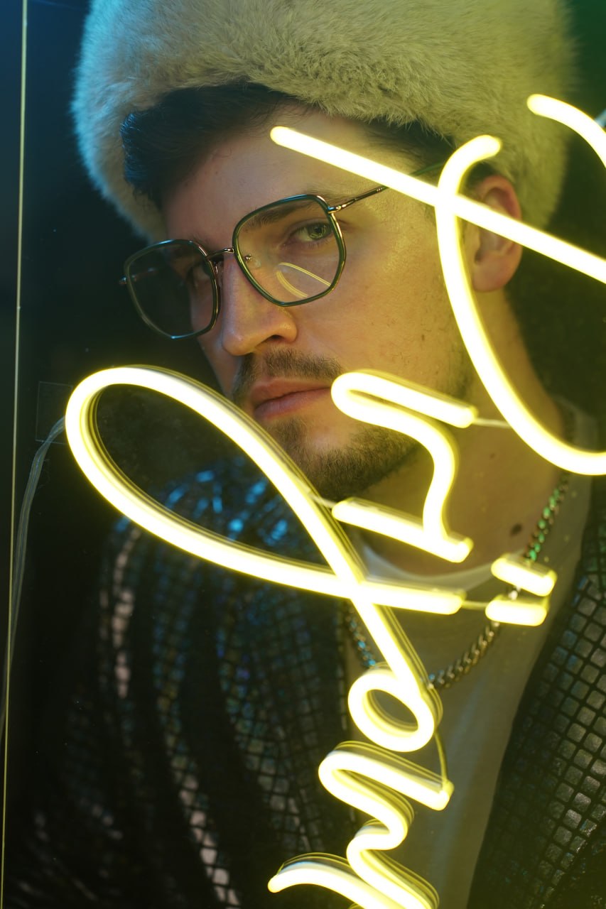 Person wearing glasses and a furry hat, illuminated by neon lights, with a serious expression.