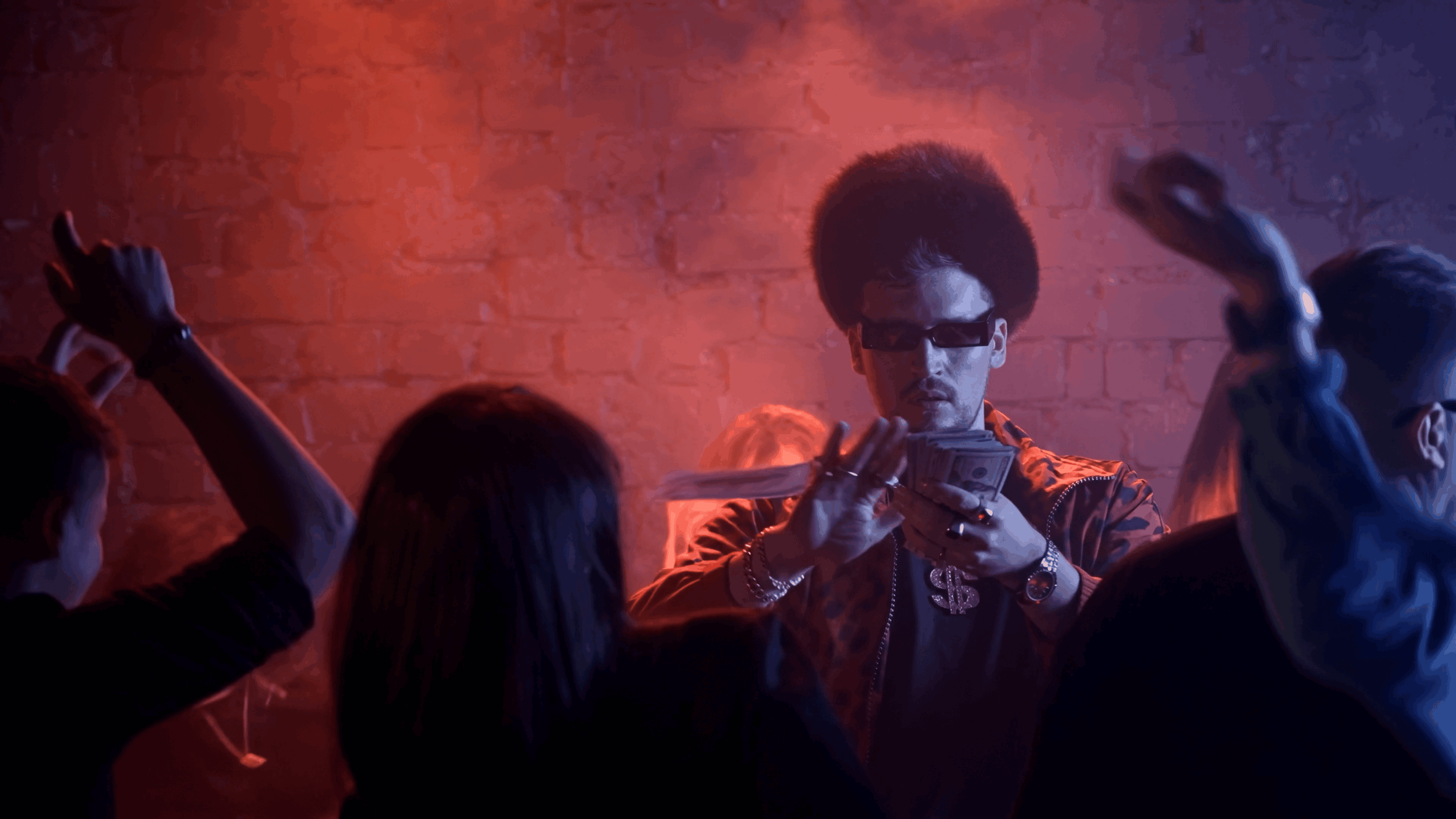 Person with sunglasses and curly hair playing a guitar surrounded by people in a dimly lit room.
