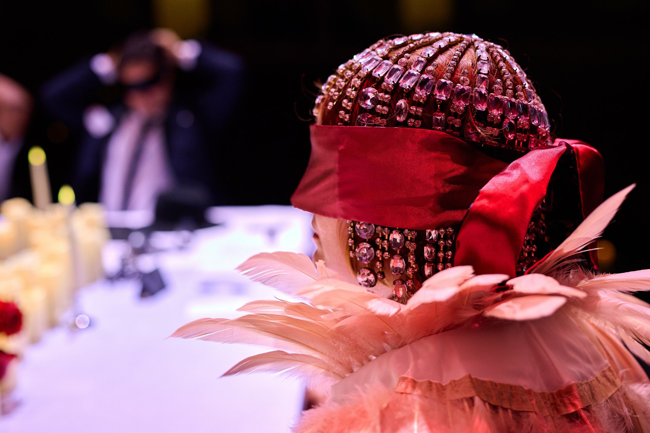 Person with elaborate head covering and red blindfold, surrounded by feathers, at a table with blurred background.
