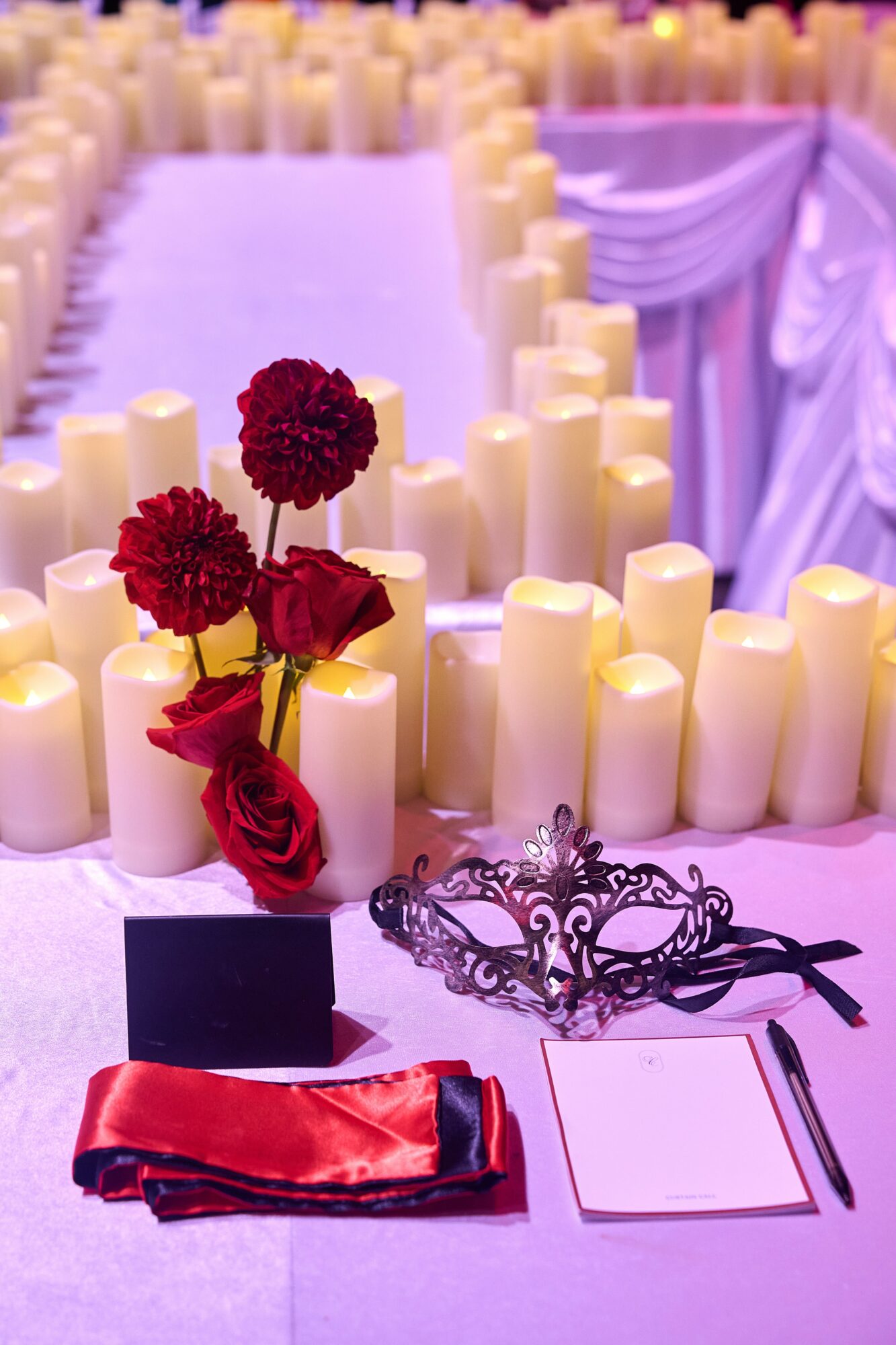 Table with red flowers, black mask, red ribbon, and notepad, decorated with candles and draped fabric in background.