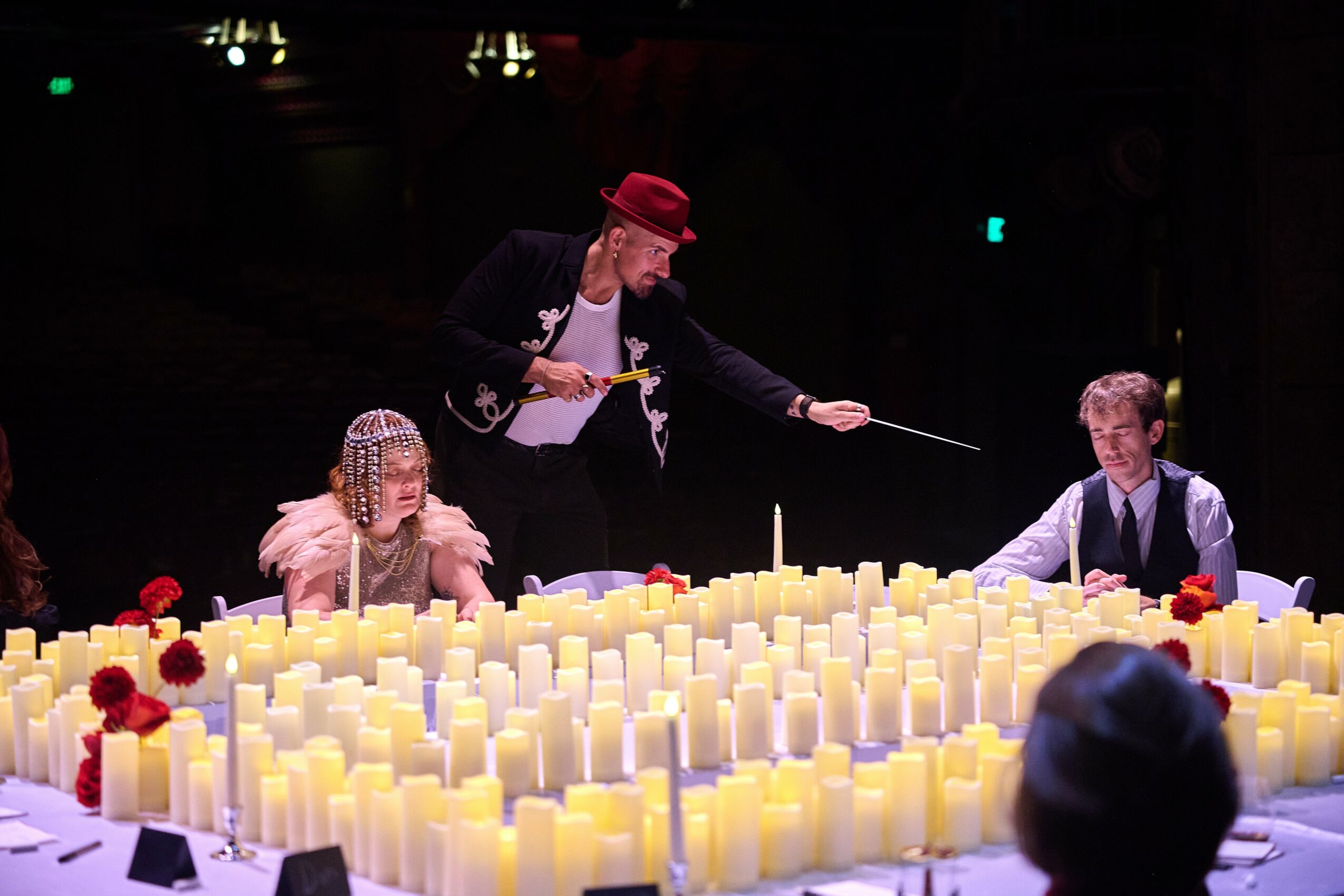 Three people sit at a table with numerous lit candles and red flowers, one person stands holding a stick, in a dark setting.