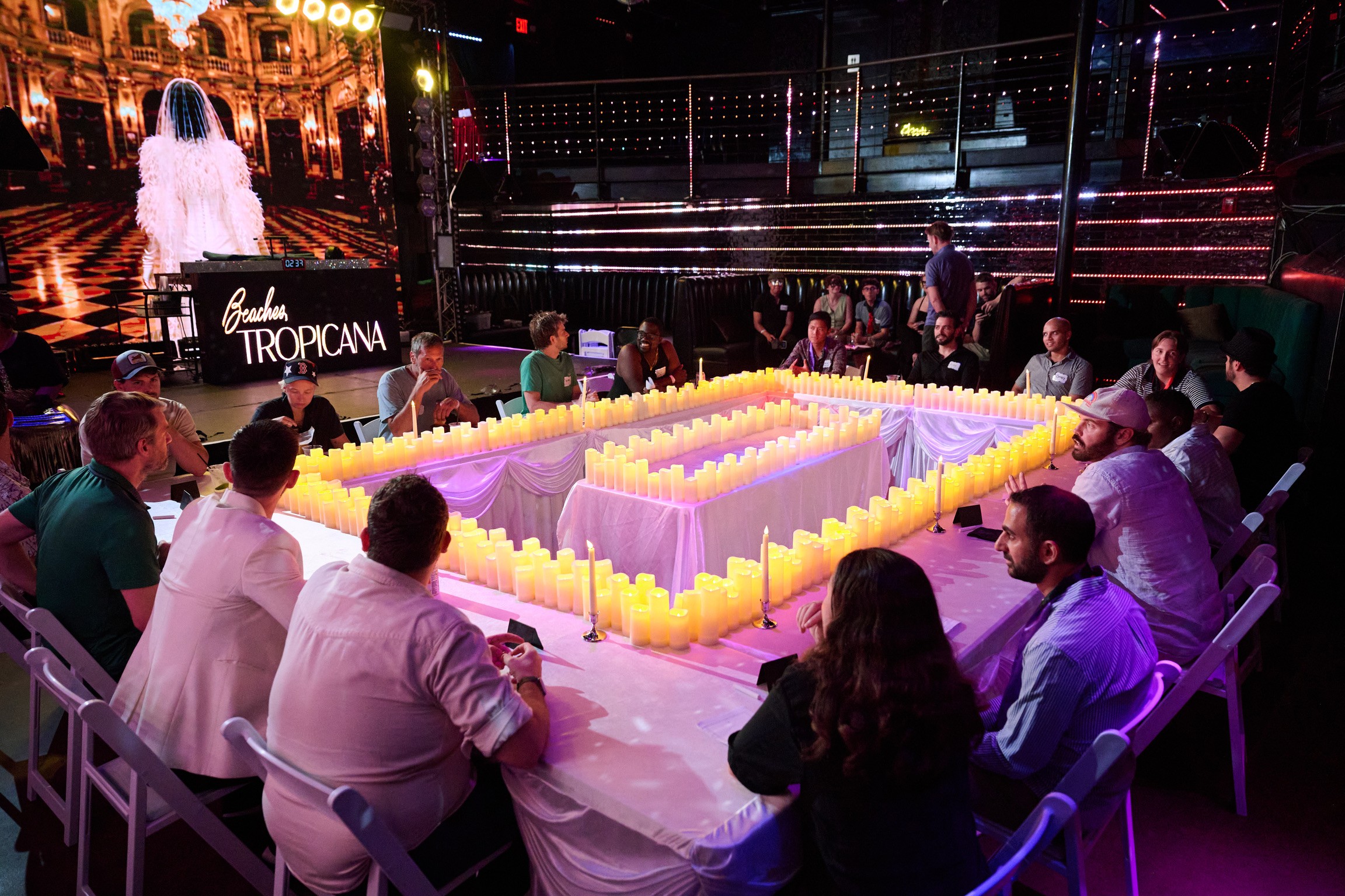 People sit around a table with a candle-lit centerpiece at a nighttime event, with a stage and screen in the background.