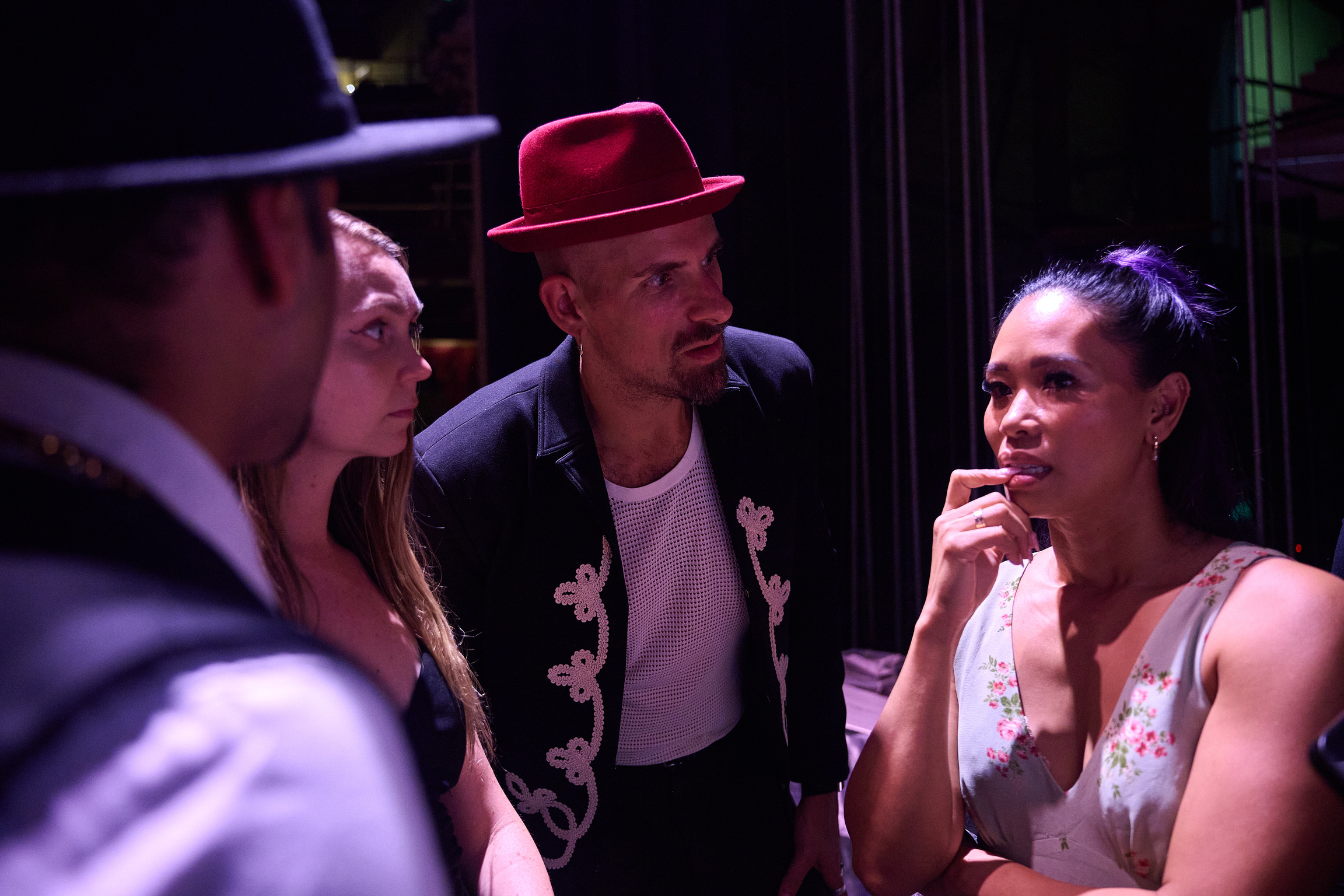Four people engaged in conversation, one woman smiling and touching her chin, others listening attentively, in a dimly lit setting.