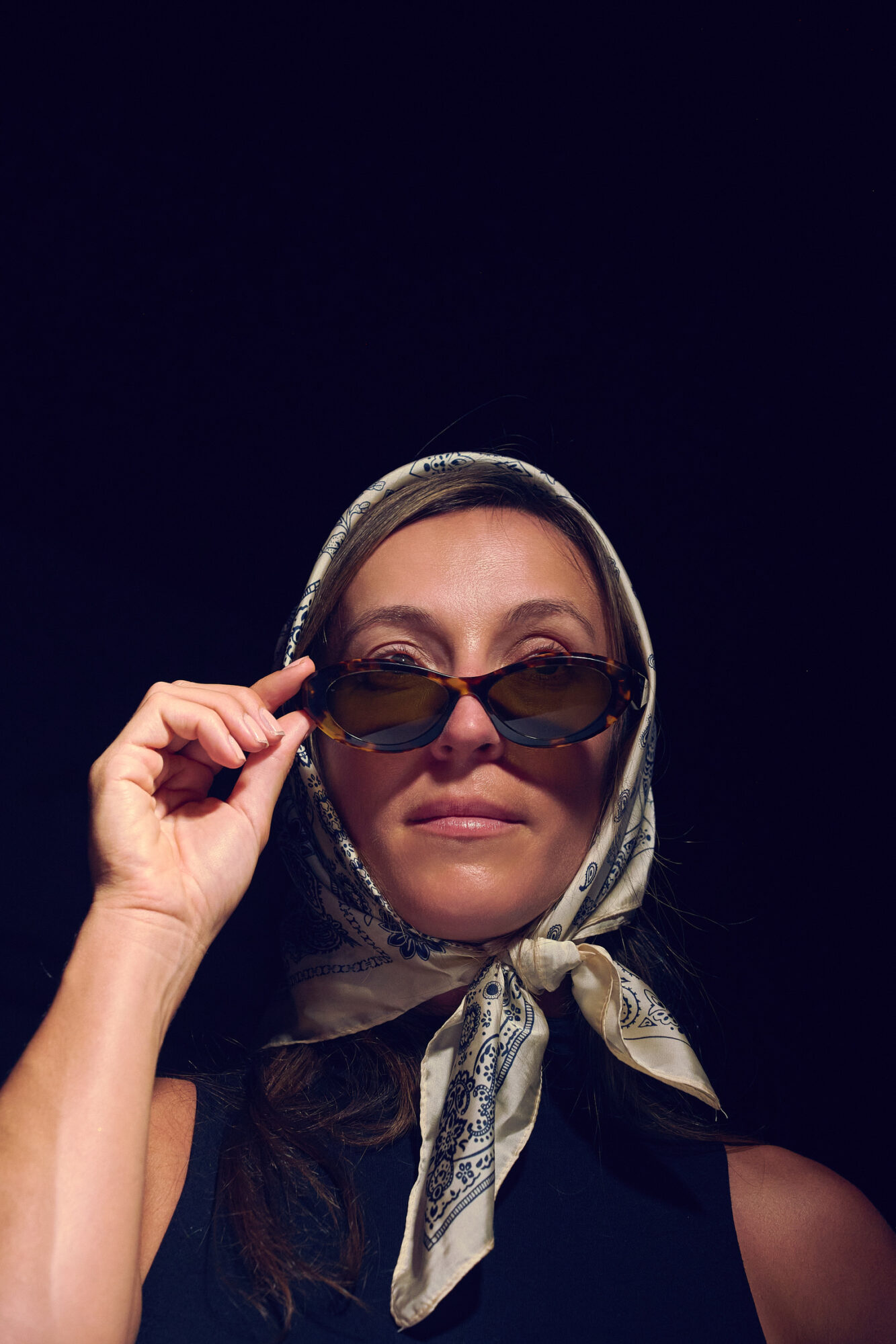 Woman with a headscarf and sunglasses looking at the camera, dark background, close-up portrait.
