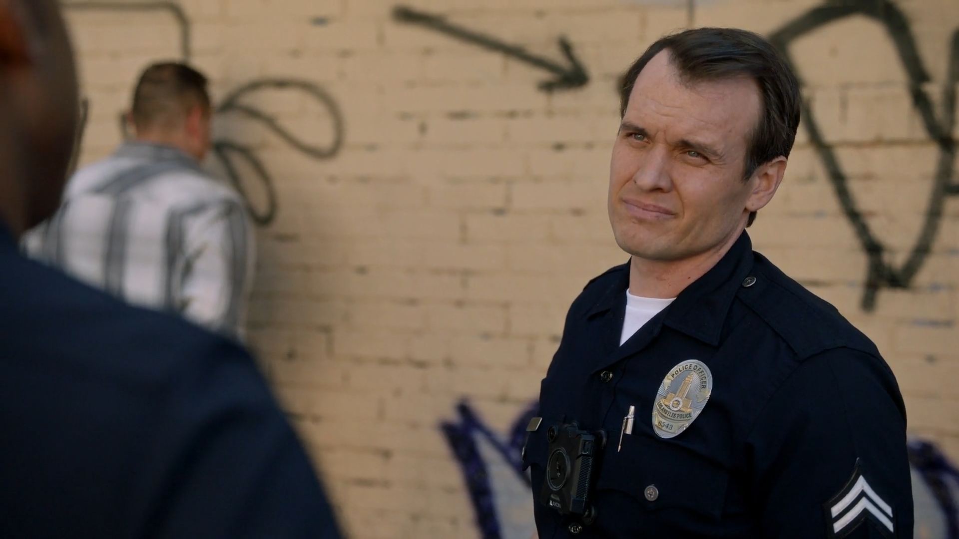 Police officer with a badge and uniform, standing outdoors against a graffiti-covered brick wall, looking to the side.