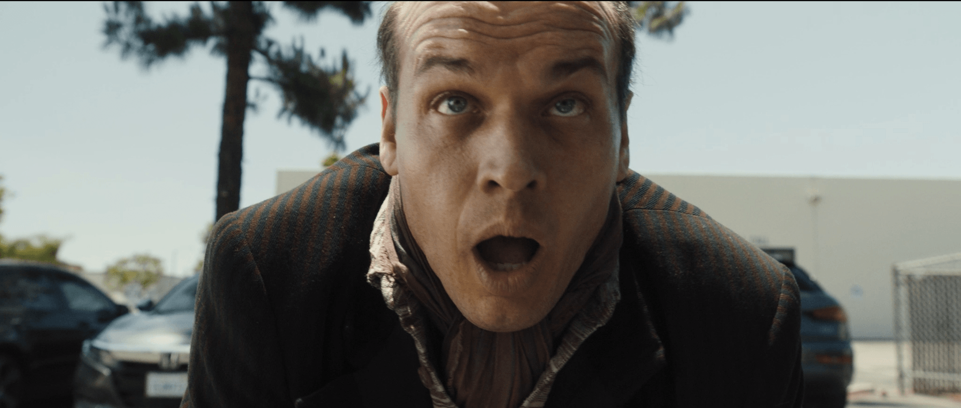Man with surprised expression leaning forward outdoors, cars and trees in background.
