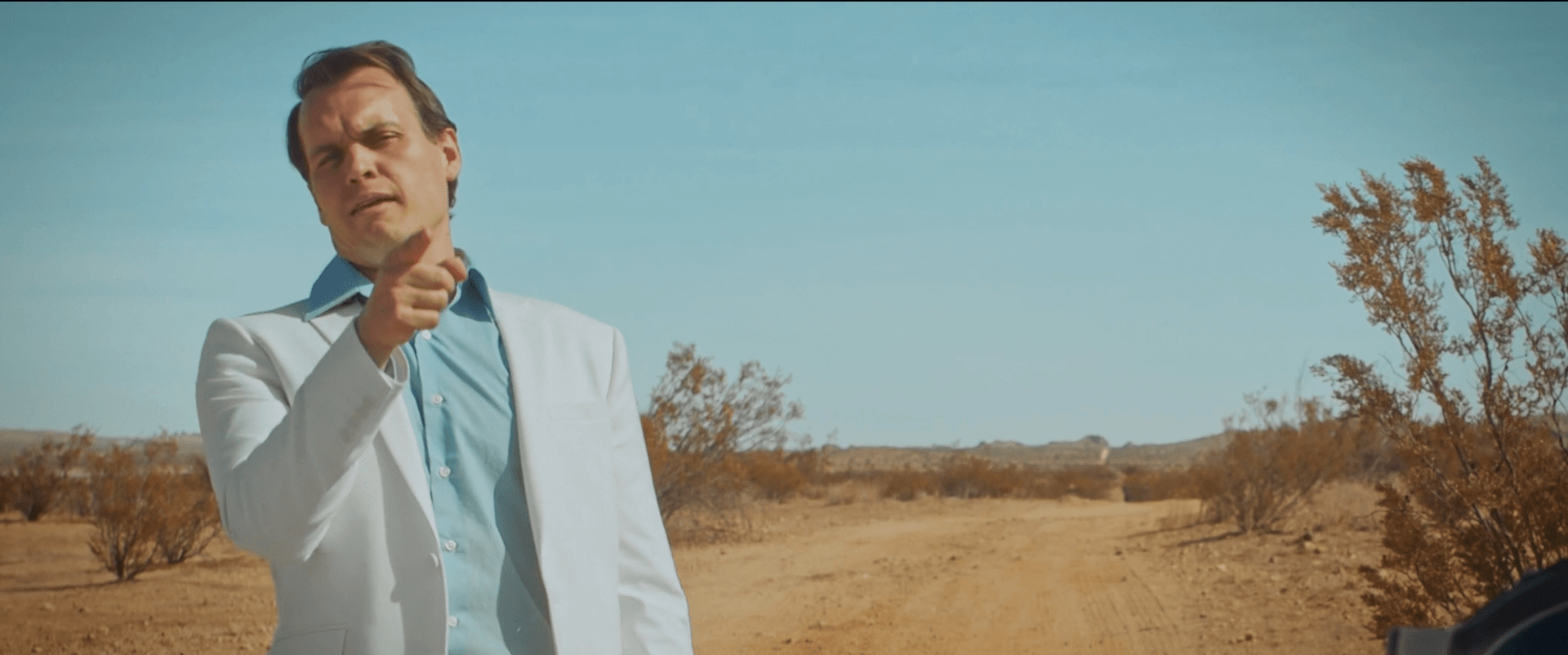 Man in a white jacket standing outdoors in a desert landscape with sparse bushes and a clear sky.