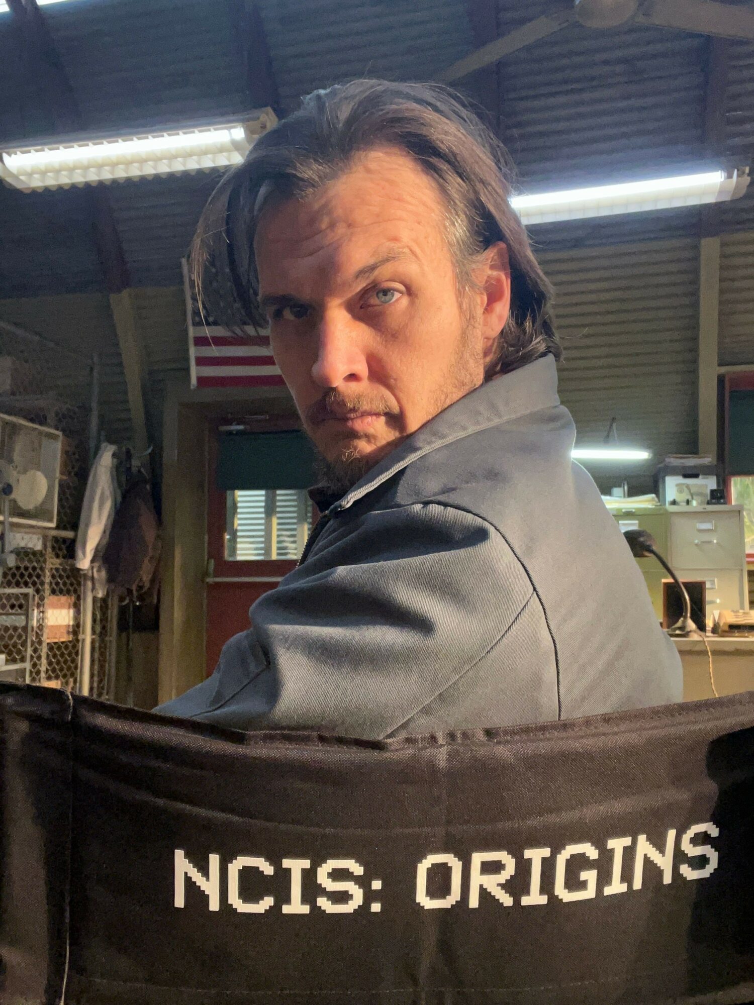 Man with long hair and beard in a gray jacket, looking sideways, behind a black NCIS: ORIGINS barrier.