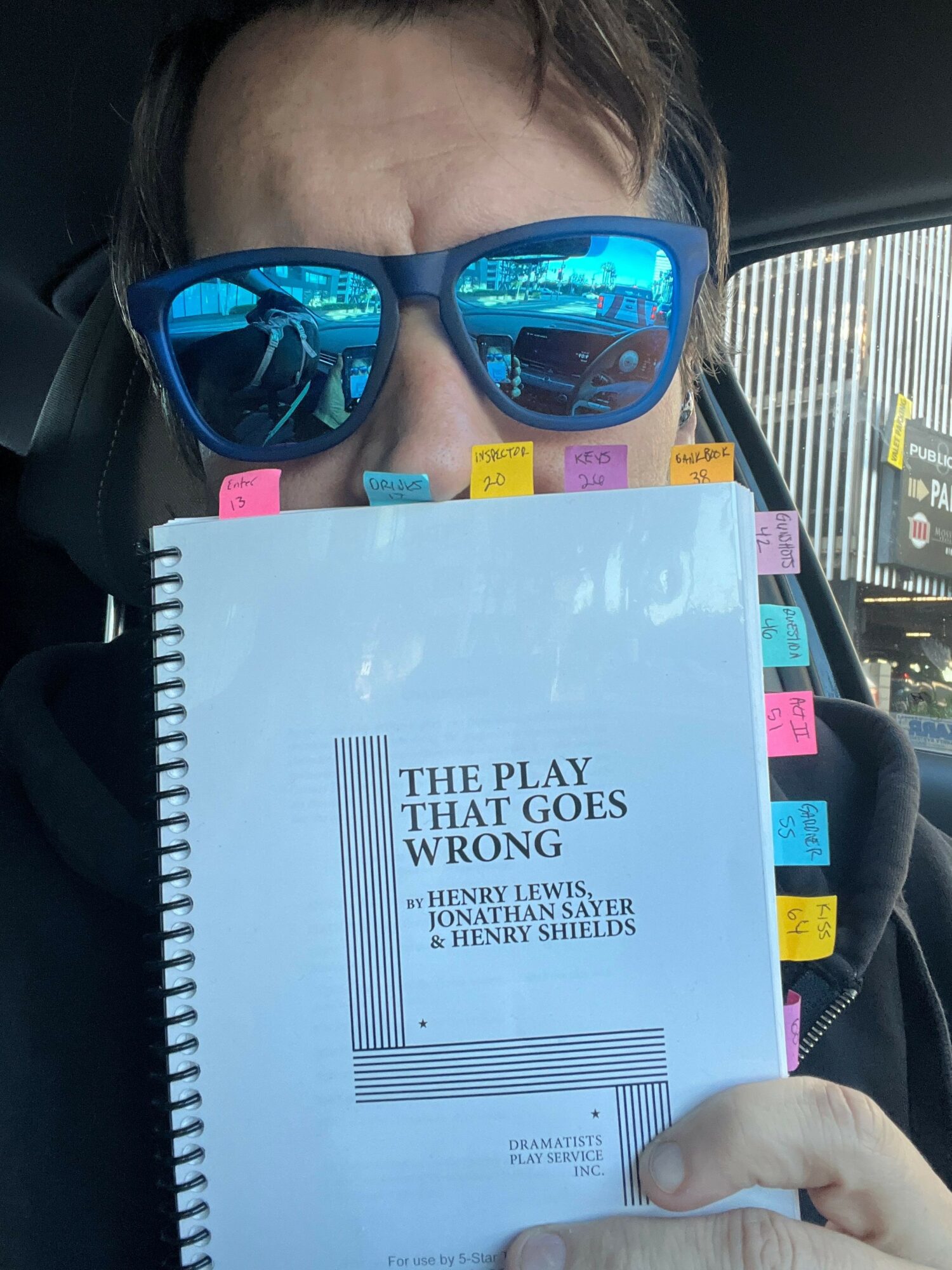 Person wearing sunglasses holding a spiral notebook with sticky notes, inside a vehicle.