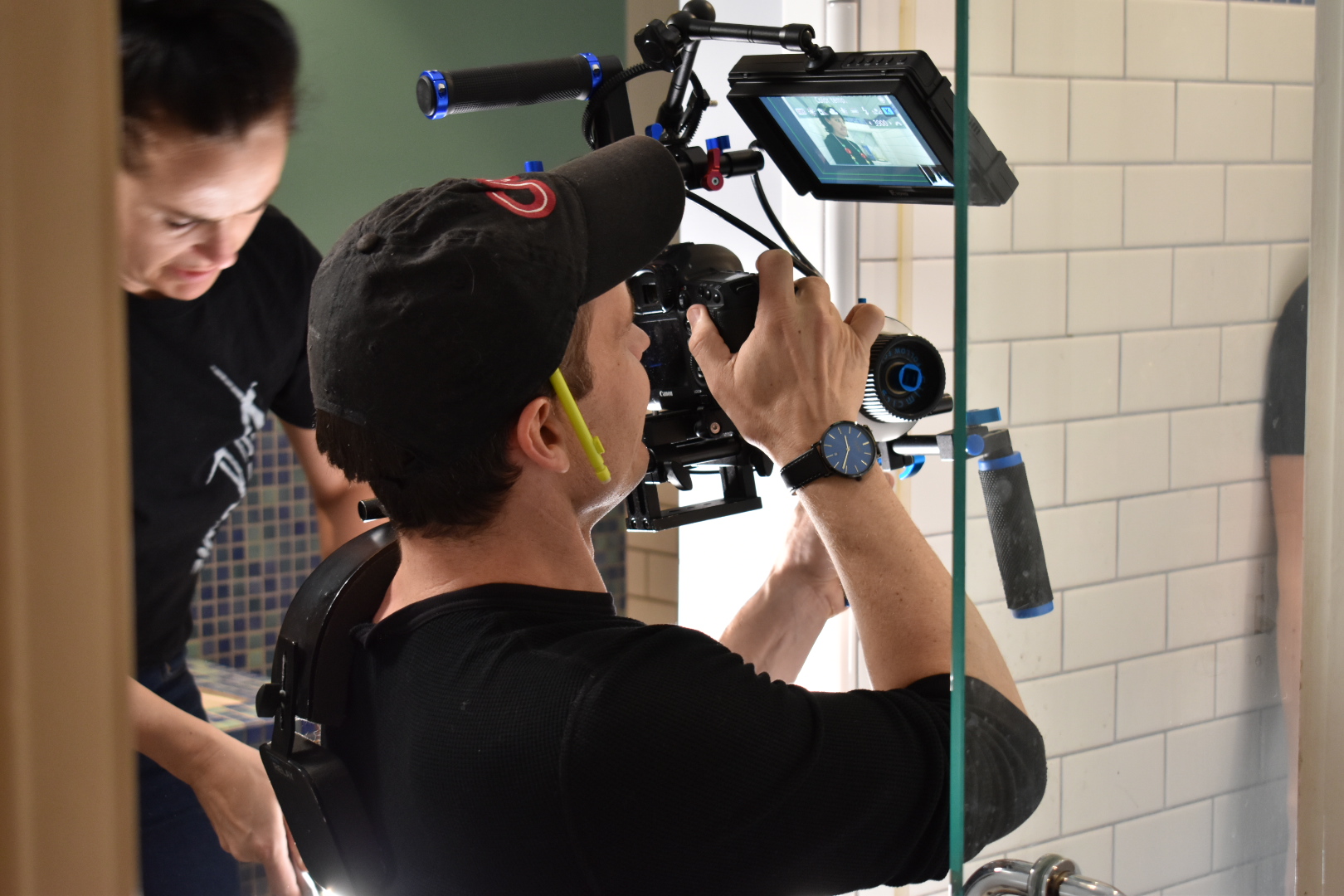 Person operating a professional camera with a monitor, with another person nearby, in an indoor setting with tiled walls.