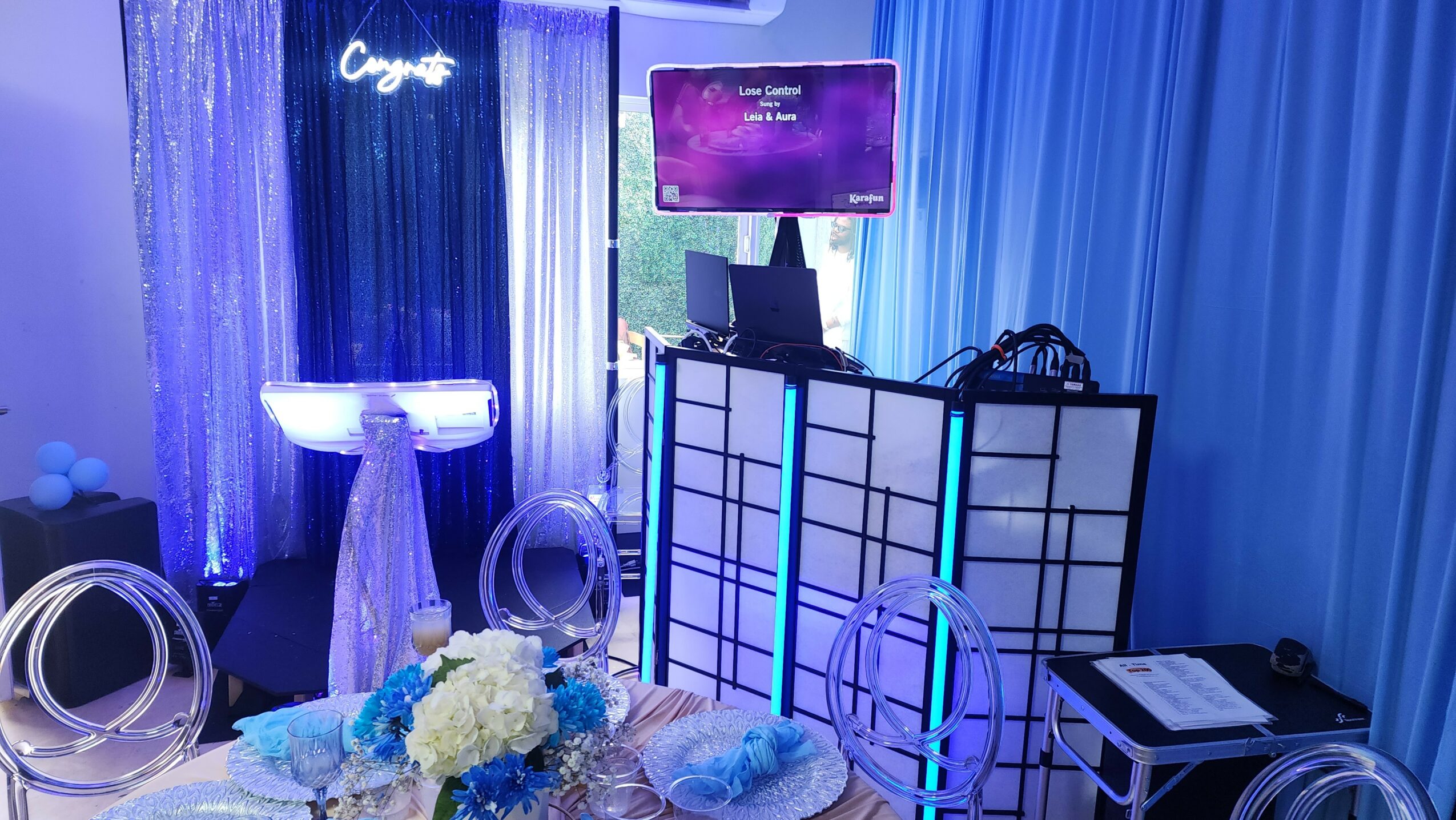 Event setup with blue lighting, a screen, tables with floral arrangements, and decorative rings, in a dimly lit room.