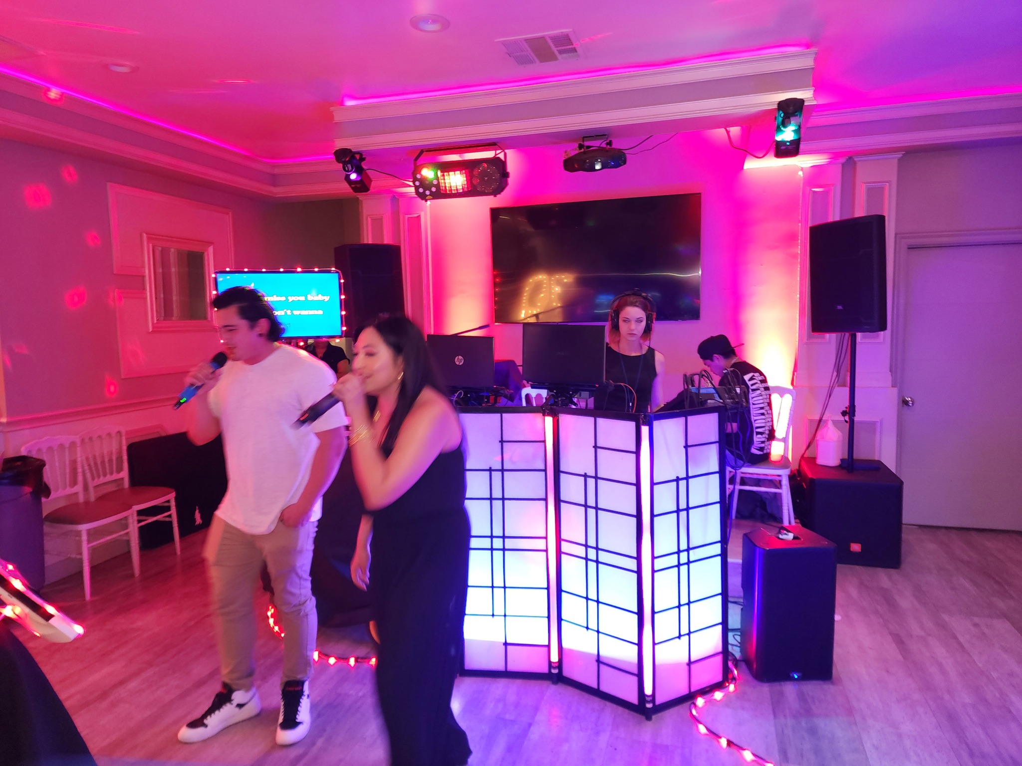 People dancing in a room with pink and purple lighting, a DJ booth, and a large screen in the background.