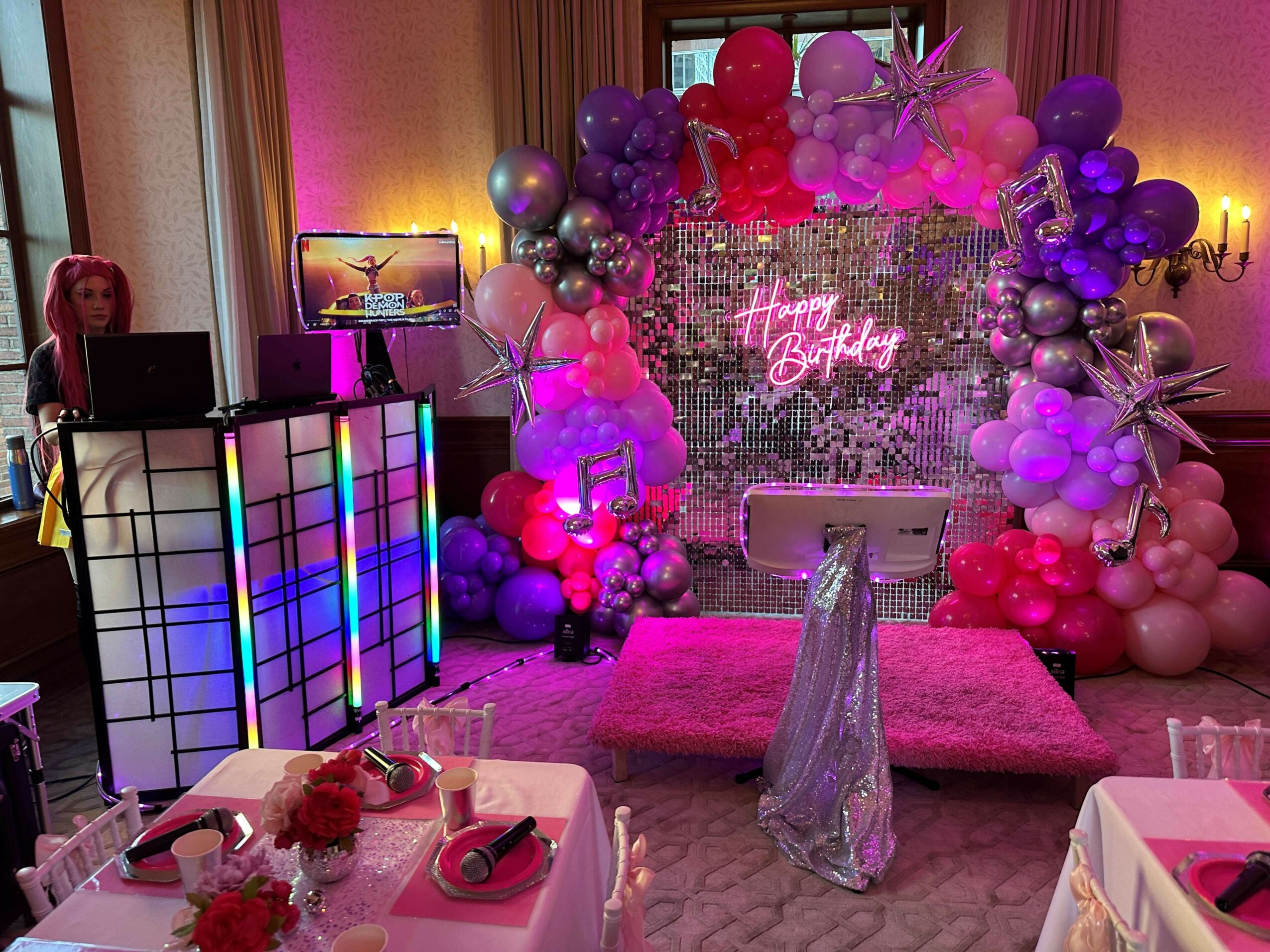 Party room with balloon arch, pink carpet, and decorated table, celebrating a birthday with colorful decorations and lighting.