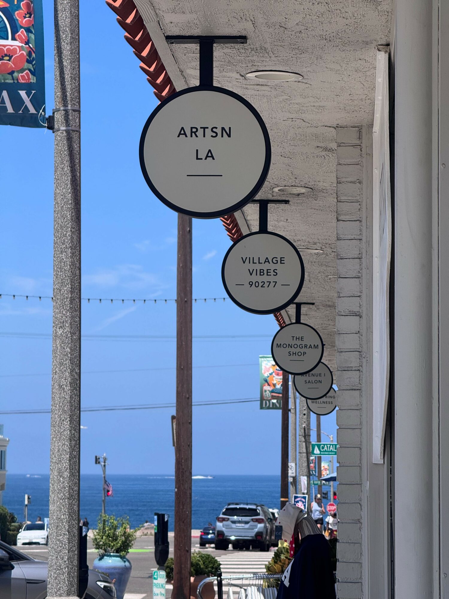 Street view with hanging circular signs, ocean in background, cars parked, and sidewalk with plants.