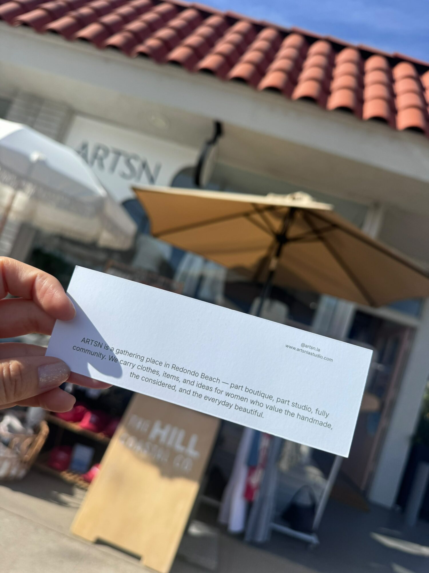 Person holding a white check in front of a storefront with a tiled roof and an umbrella, partially obscured sign reading 'ARTISAN'.