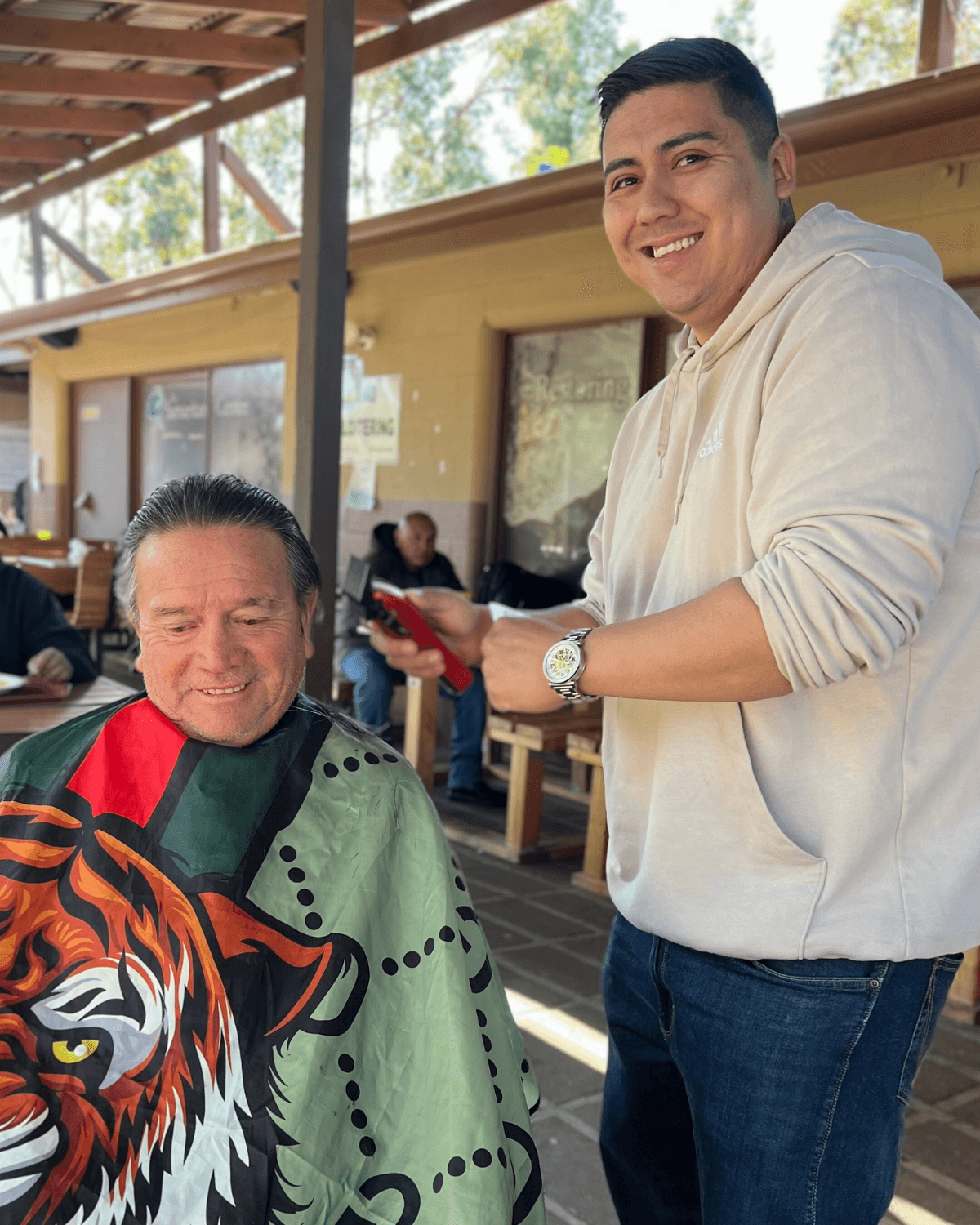 Two men outdoors, one smiling and seated with a tiger graphic on his shirt, the other standing and holding a phone.
