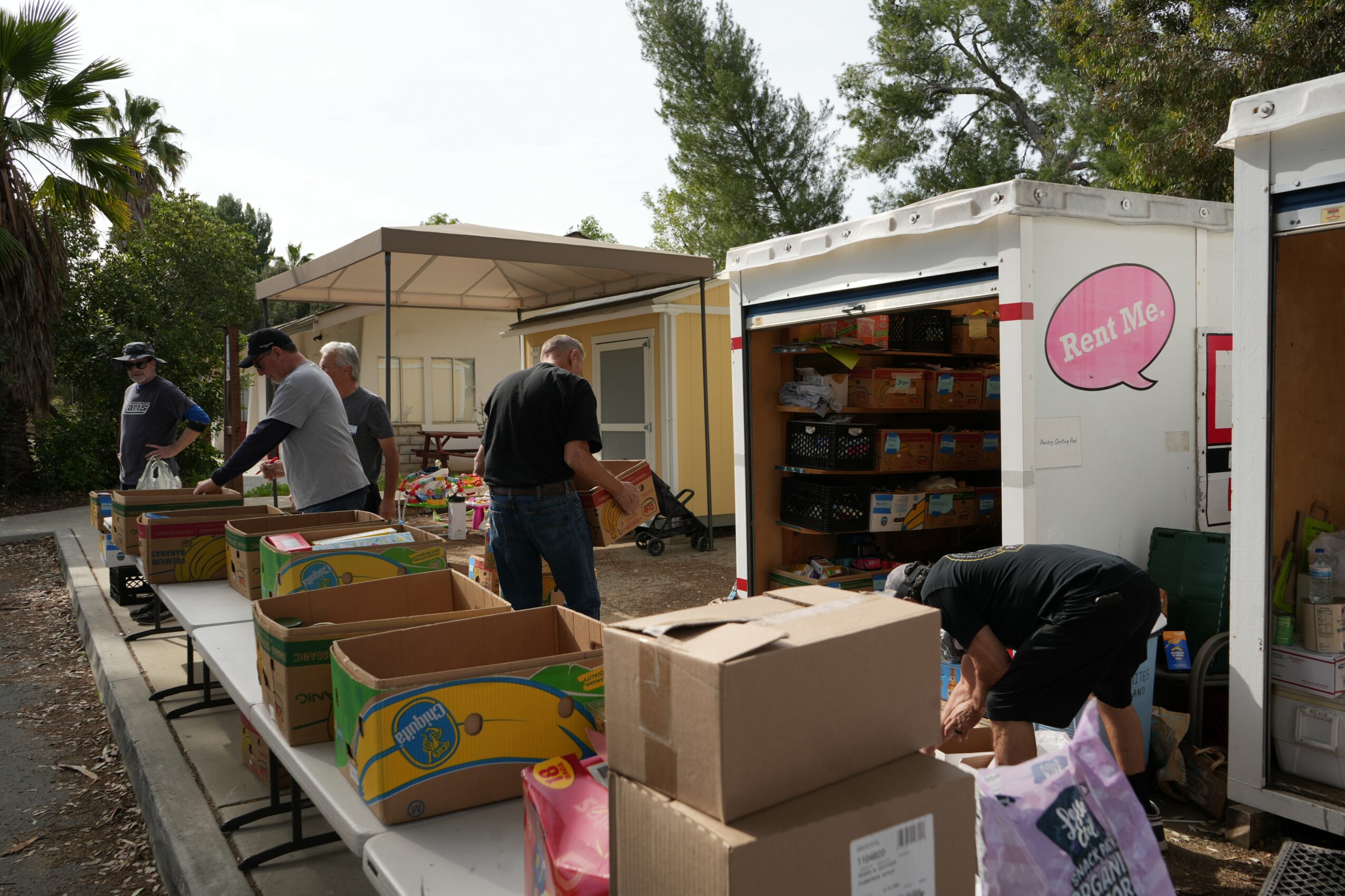 People browsing tables with boxes and a mobile bookshop outdoors, trees and a building in the background.