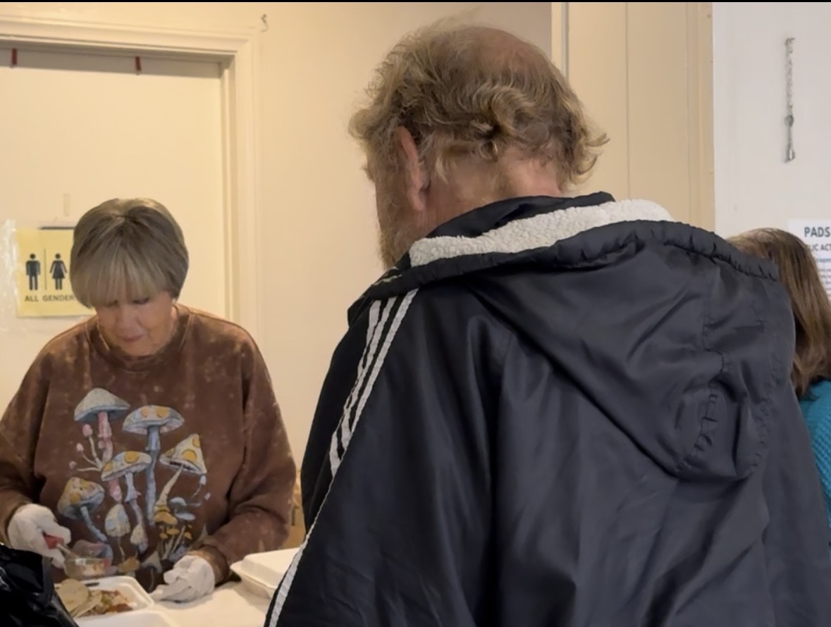 Person with curly hair wearing a dark jacket facing a woman with short gray hair, in a room with a door and a sign, possibly in a waiting area.
