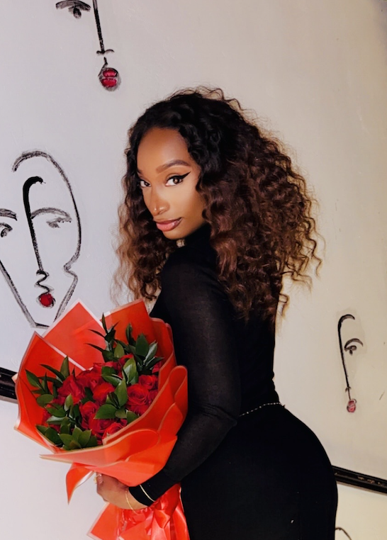 Woman with curly hair holding a bouquet of red roses, standing indoors with abstract face drawings on the wall.