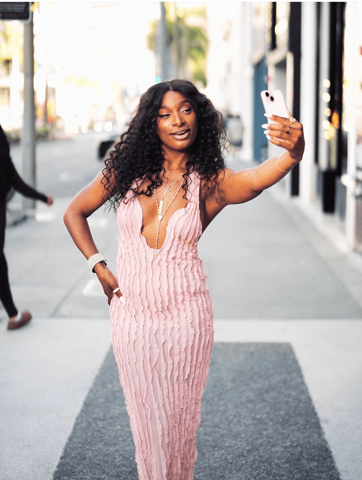 Woman with curly hair in pink dress taking a selfie on city sidewalk.