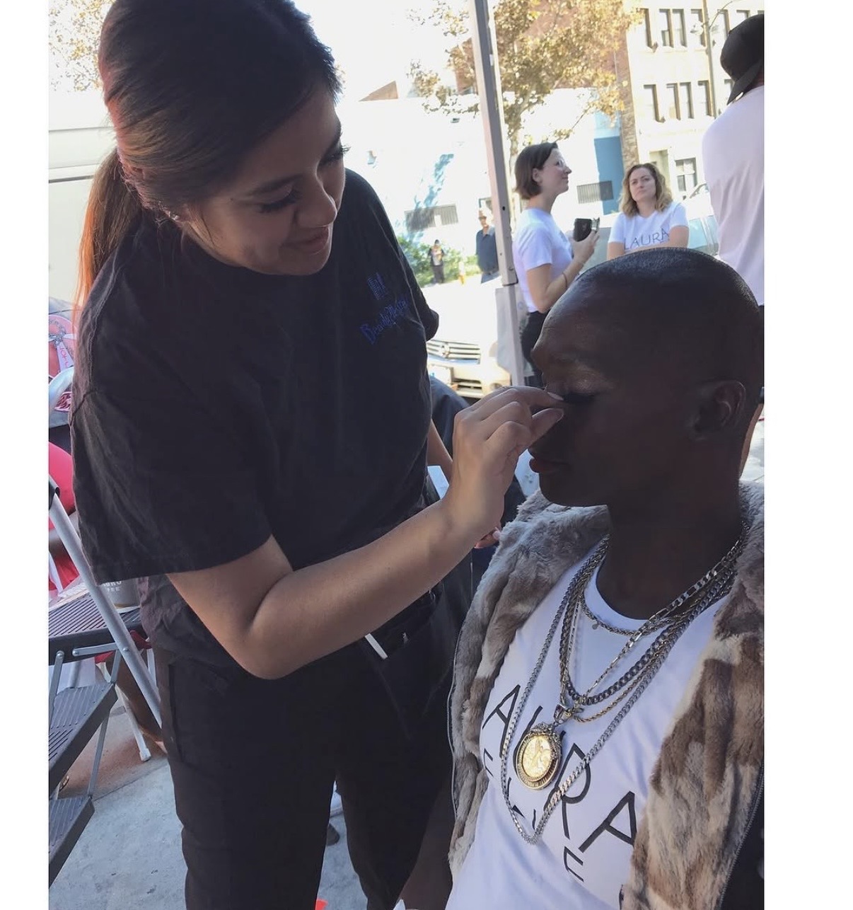 A woman applies makeup to a man with jewelry, in a setting with people in the background outside a window.