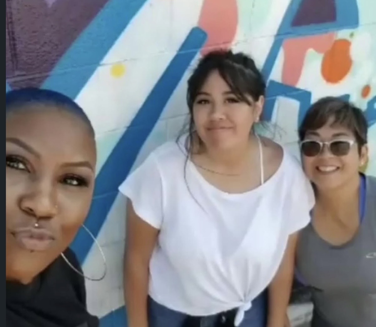 Three women standing in front of a colorful mural, smiling and posing for a photo.