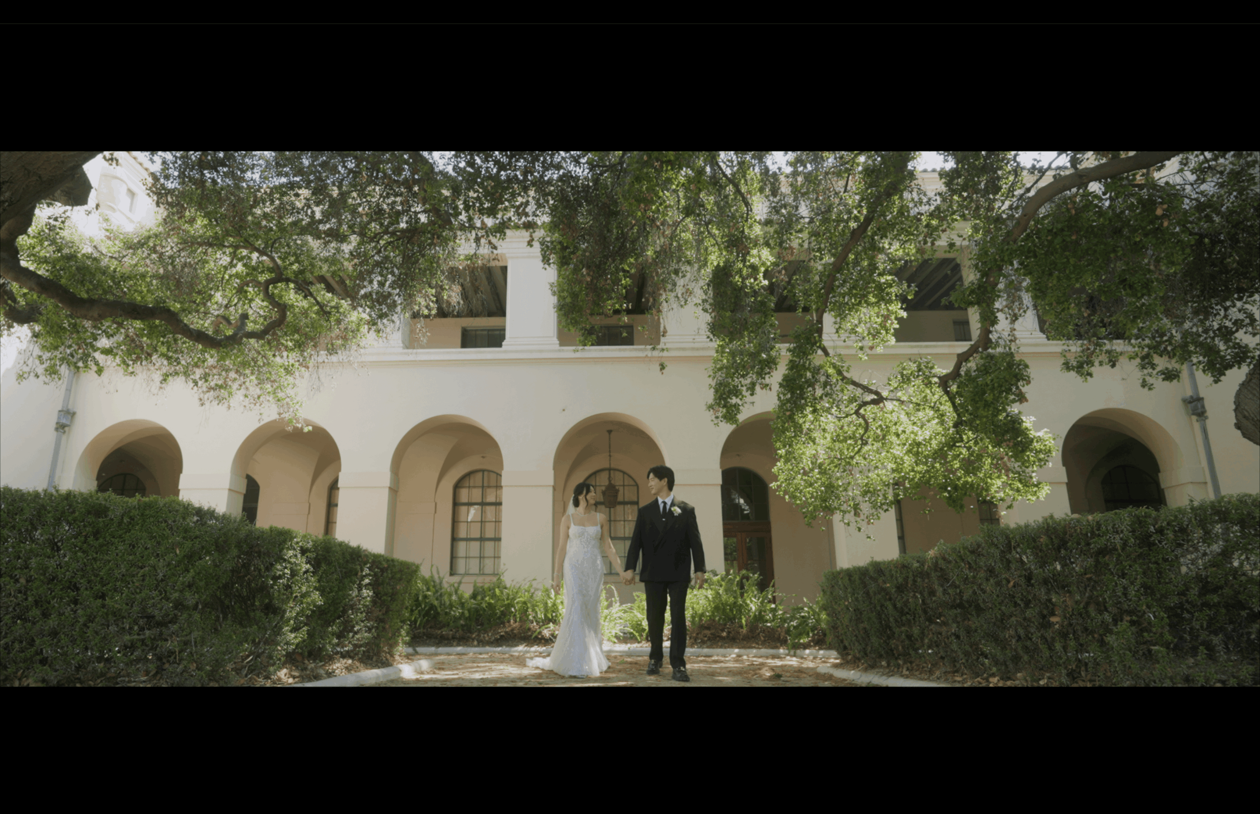 A bride and groom holding hands stand in front of a white building with arches, surrounded by greenery.