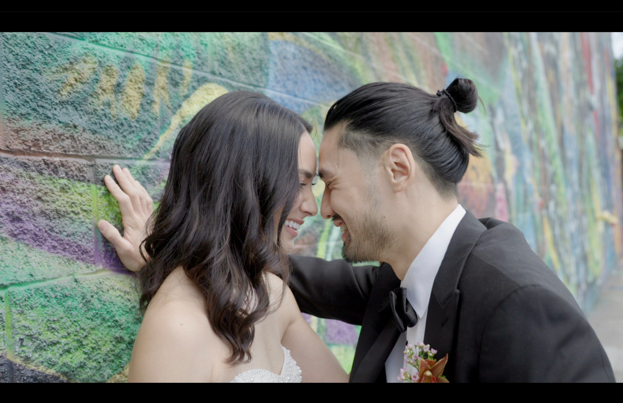 A couple with foreheads touching, smiling, close to each other against a colorful graffiti wall.