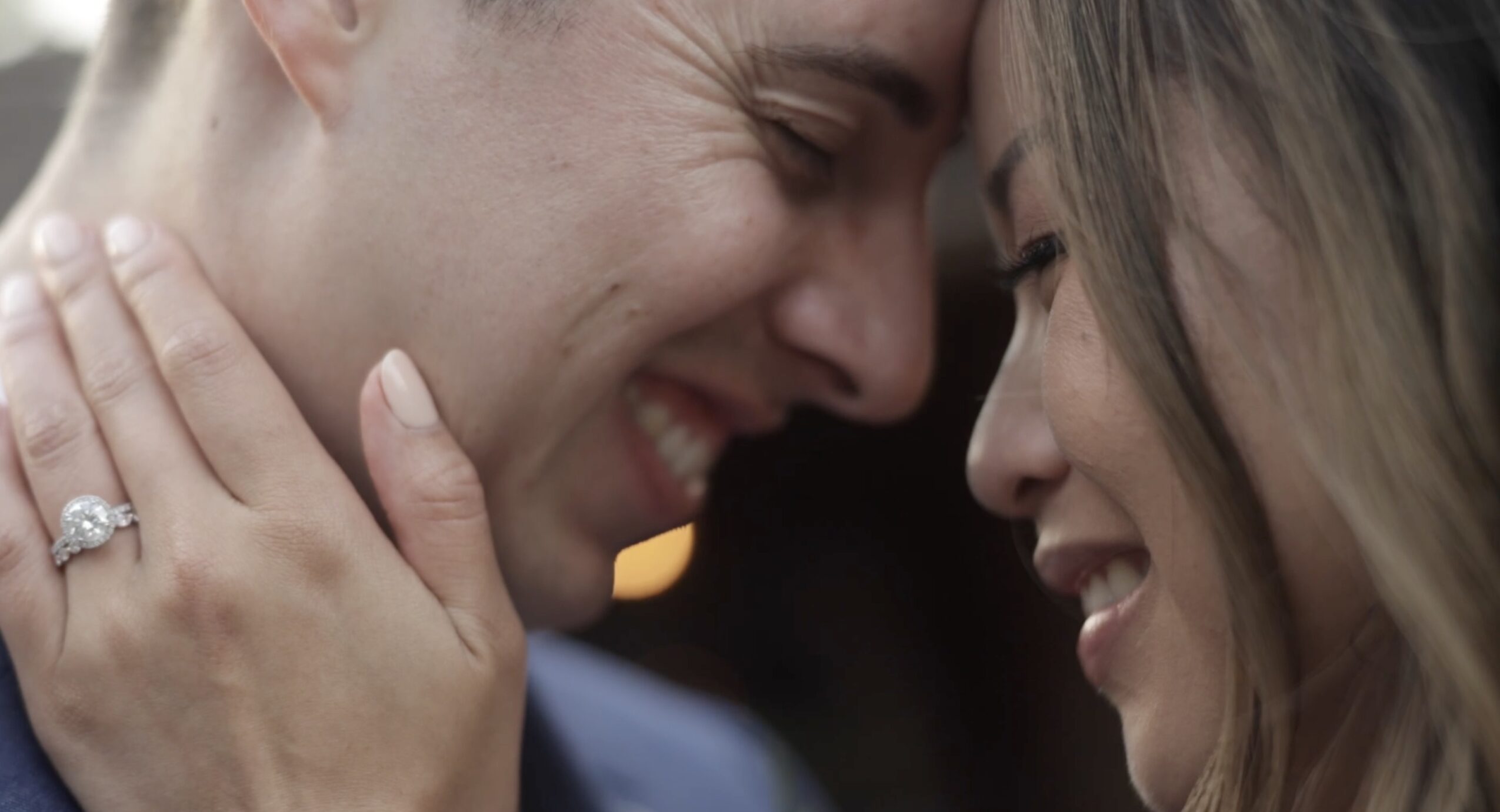 Two people close together, smiling and touching foreheads, with one wearing a ring on their finger.