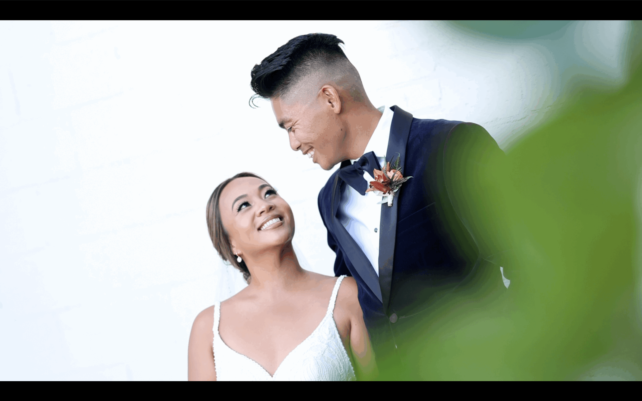 A smiling couple dressed in wedding attire looking at each other, with a blurred green plant in the foreground.