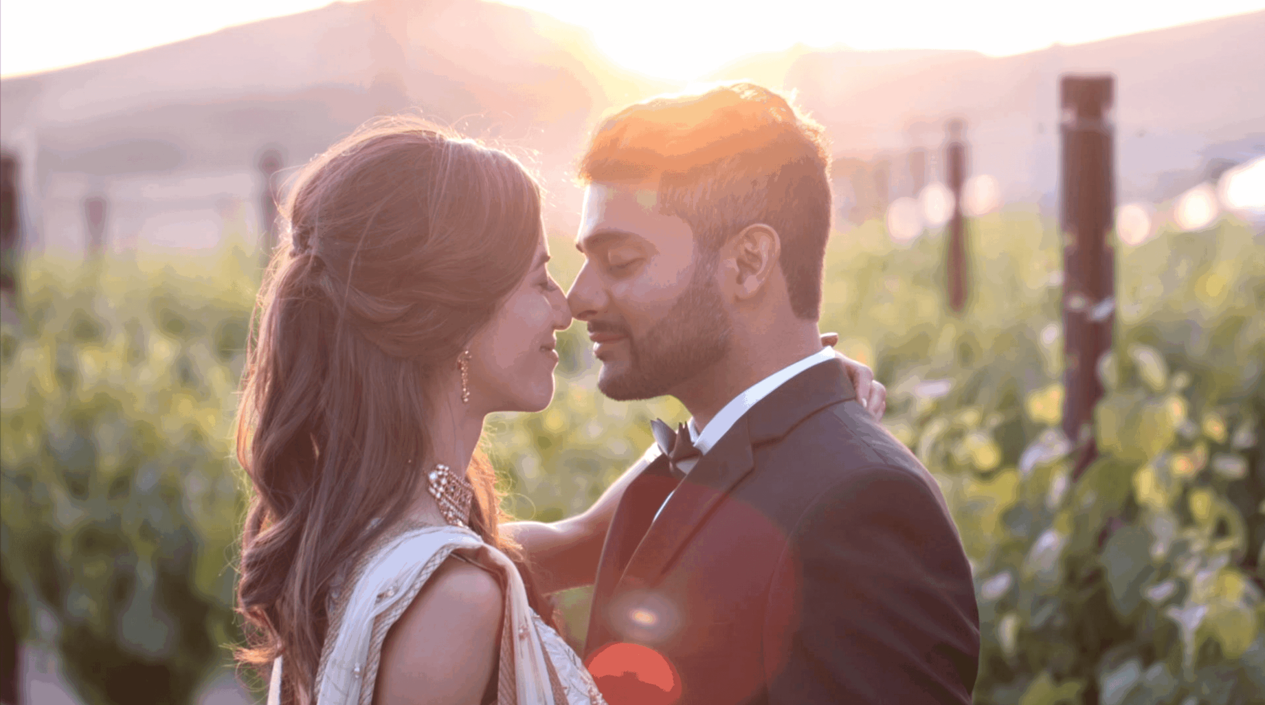 A couple faces each other closely outdoors with sunlight behind them, surrounded by greenery and a fence.