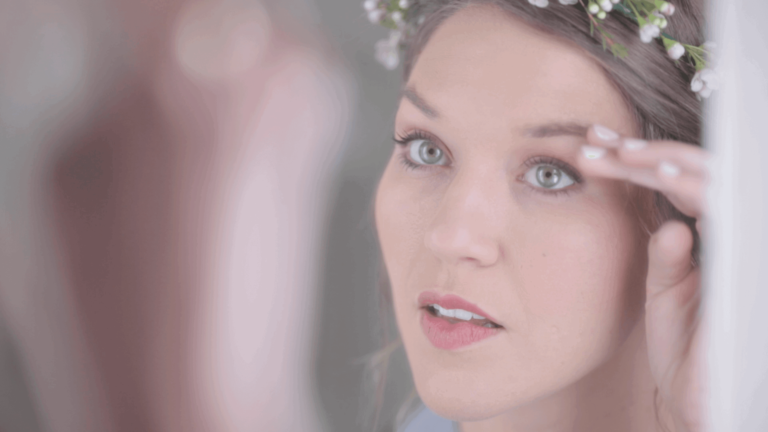 Woman with floral headband applying makeup to her eyebrow, looking into a mirror.