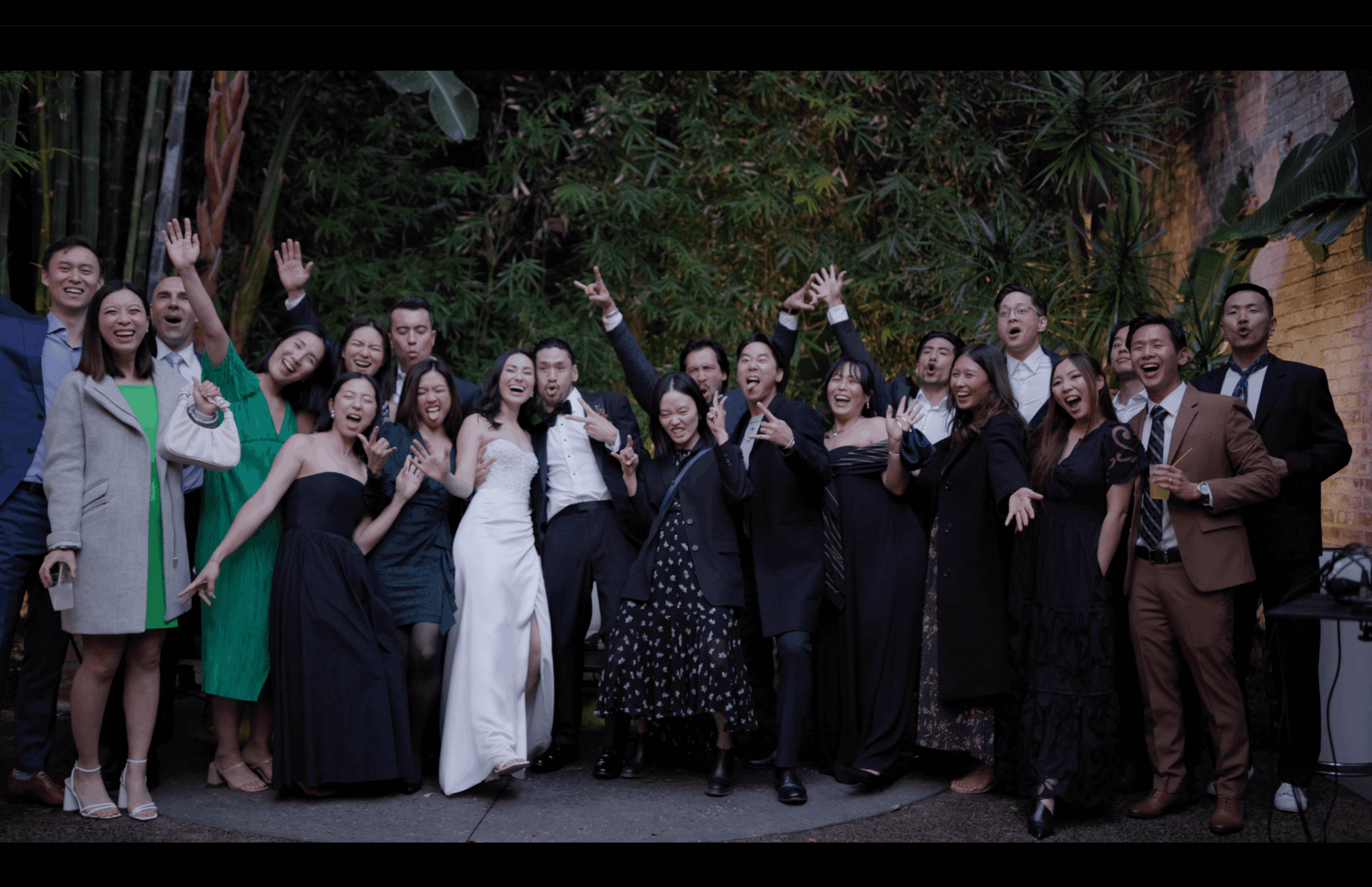 Group of people dressed in formal attire, smiling and waving outdoors with trees in the background.
