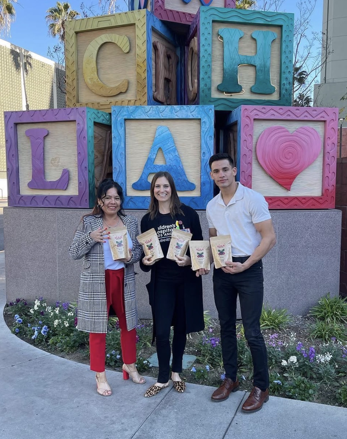 Three people standing in front of colorful blocks spelling 'CHA' with a pink heart, holding popcorn boxes, smiling outdoors.