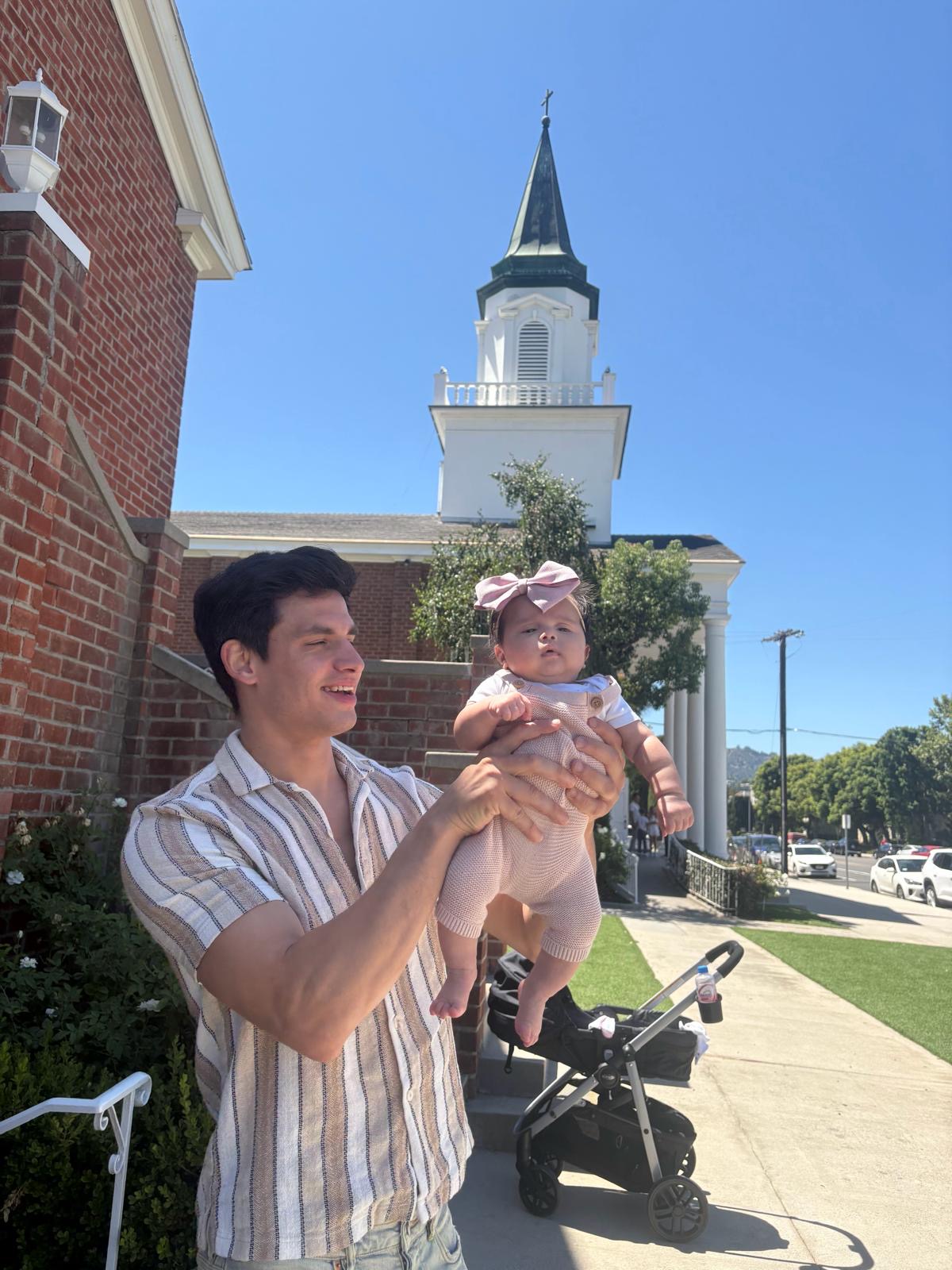 Young man holding a baby girl outdoors near a church with a tall steeple, blue sky, and sidewalk.