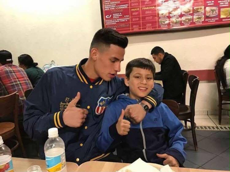 Two boys sitting at a table in a restaurant, one giving a thumbs-up, with a menu on the wall behind them.