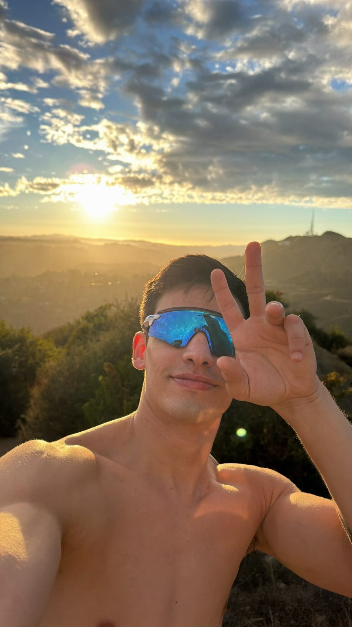 Young man taking a selfie outdoors during sunset, wearing reflective sunglasses, with a landscape and cloudy sky in the background.