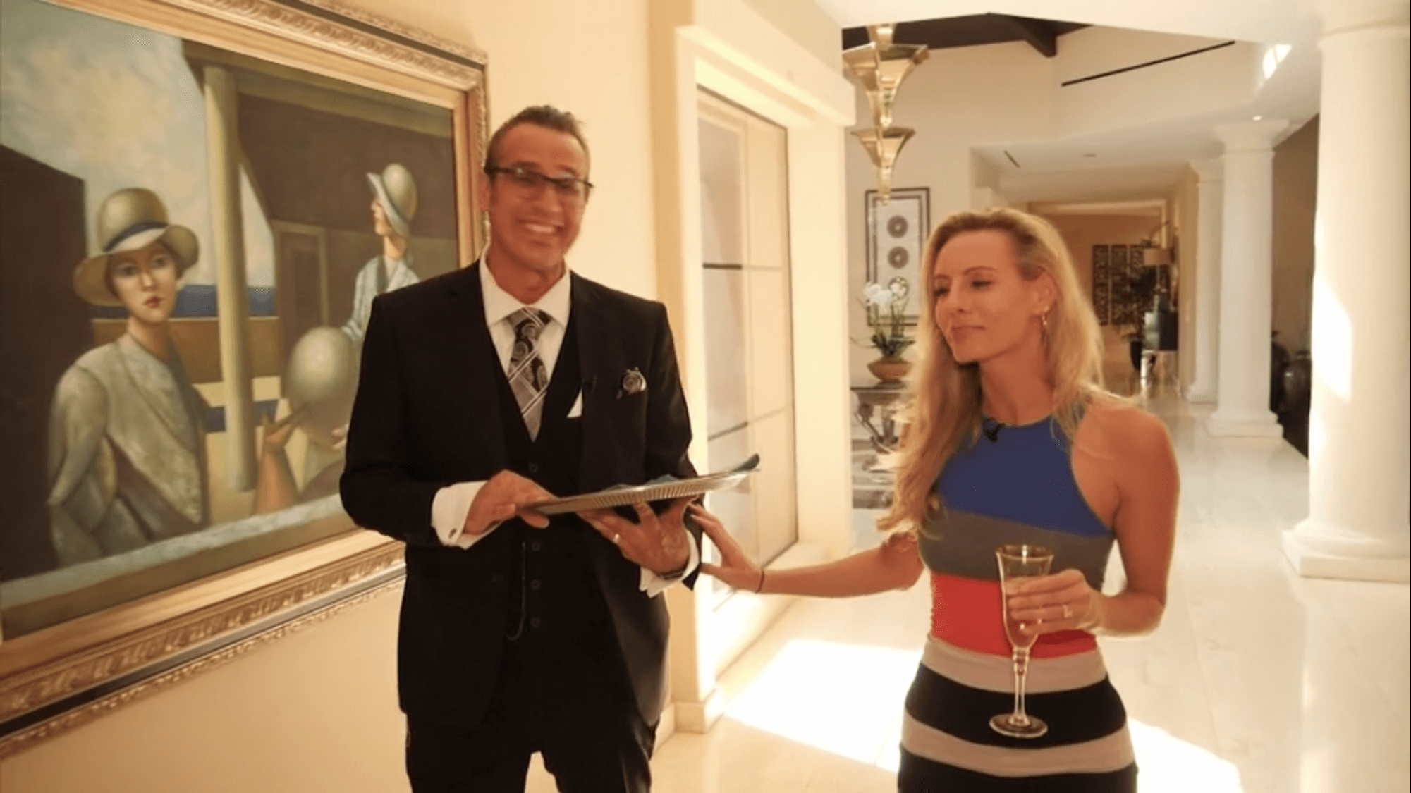 Man in a suit and woman in a dress holding a glass of champagne in a decorated hallway.