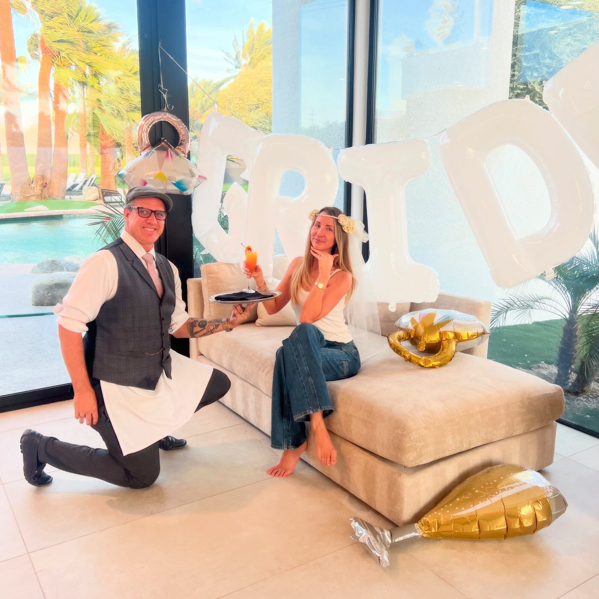 Man kneeling and woman sitting on a beige sofa with large white letters behind them, celebrating a birthday with balloons and decorations.