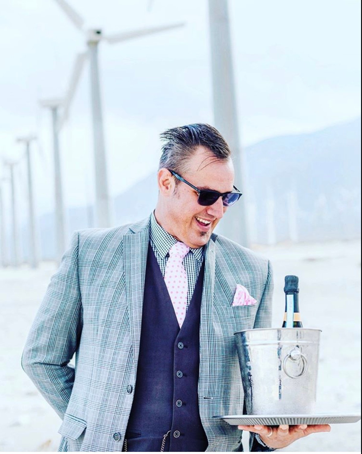 Man in suit and sunglasses holding a laptop with a microphone in an outdoor setting with wind turbines in the background.