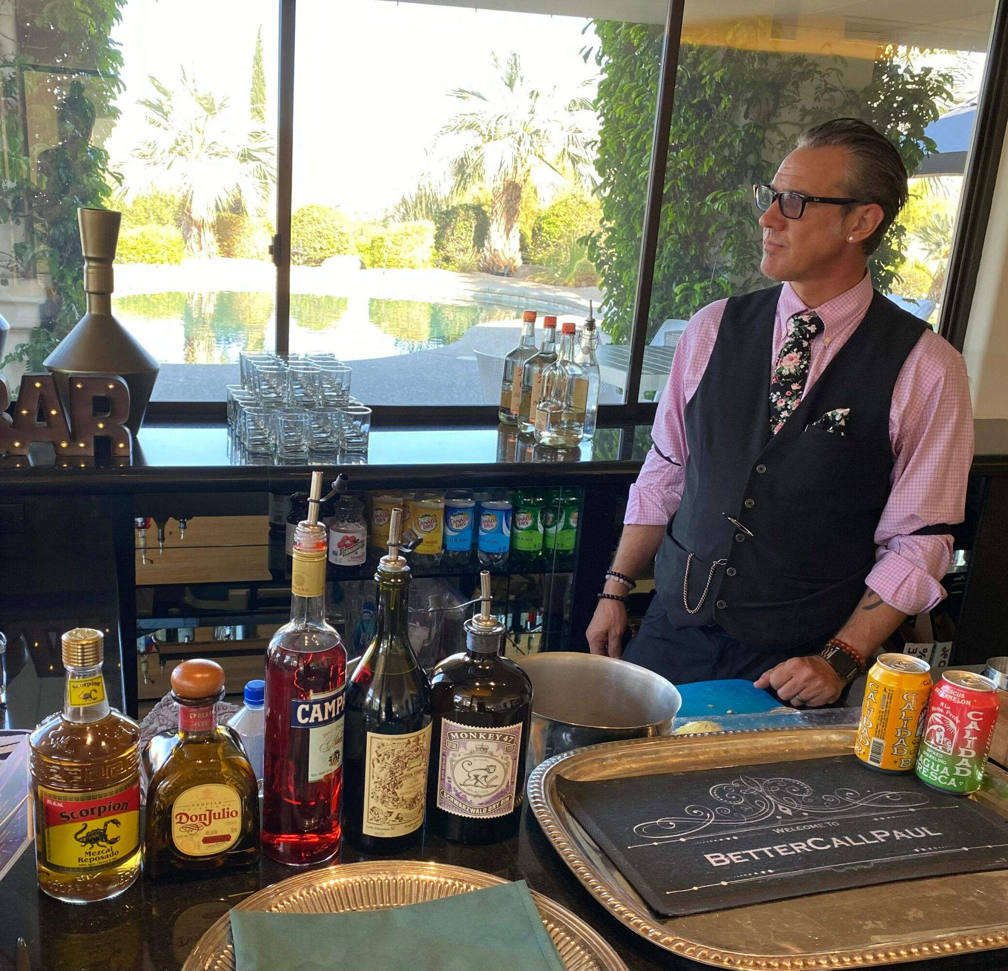 Man in vest and glasses standing behind bar with bottles and snacks, large window with outdoor view in background.