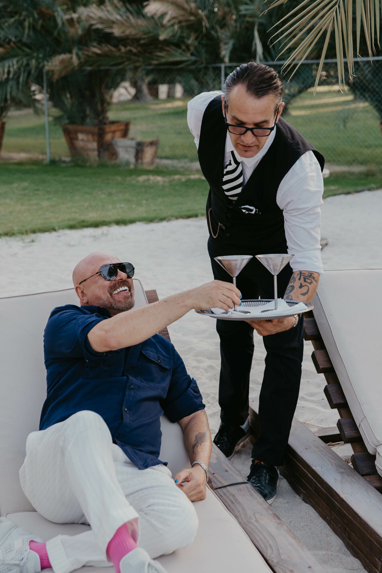Person sitting outdoors with sunglasses, smiling, being served drinks by a waiter holding a tray, in a garden setting.