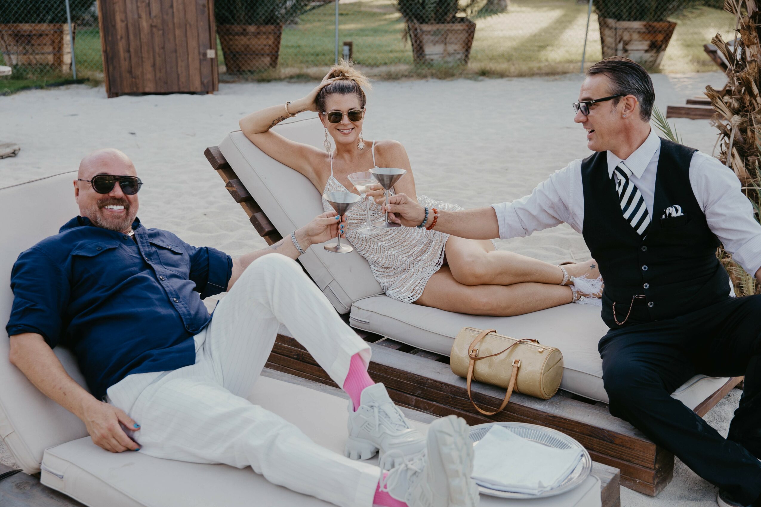 Three people relaxing outdoors, two men and a woman, with the woman lying on a lounge chair holding a drink, and the men sitting nearby, one with a drink, smiling and engaging.