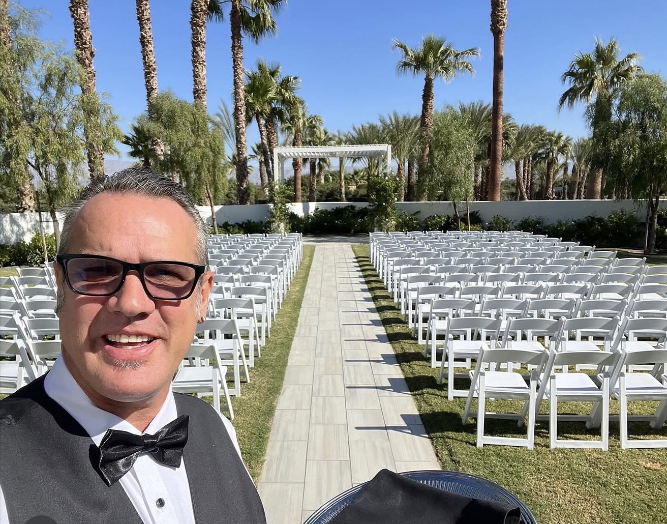 Man in tuxedo and glasses smiling outdoors with rows of white chairs and palm trees in background.