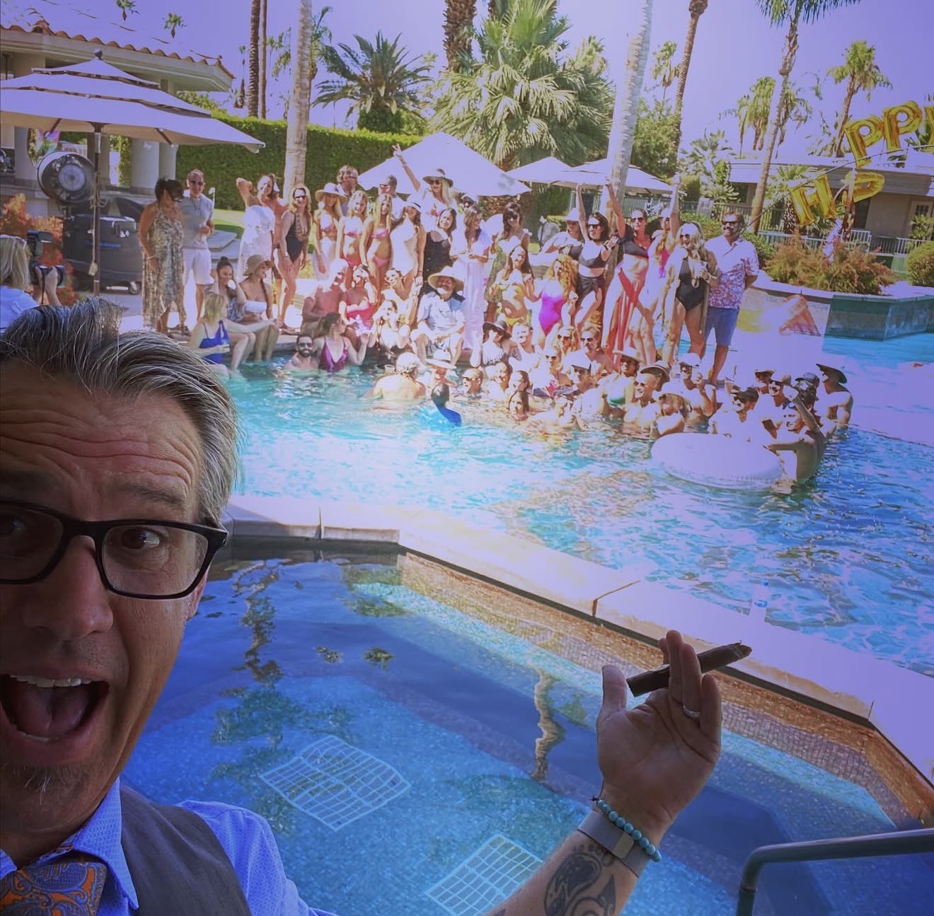 Person taking a selfie at a pool party with many people, palm trees, and umbrellas in the background.