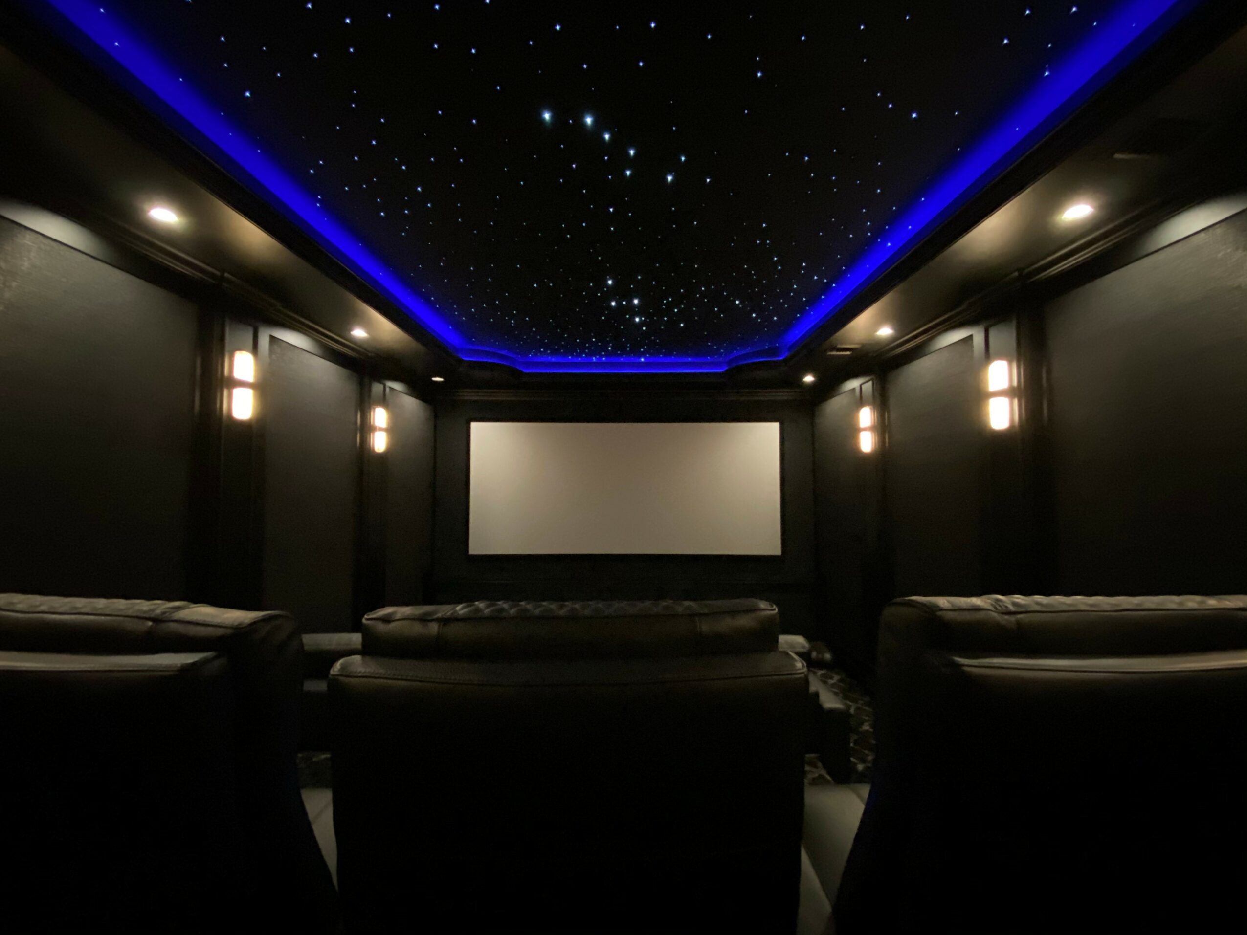 Home theater room with a large screen, black walls, and starry ceiling with blue lighting, and comfortable seating.