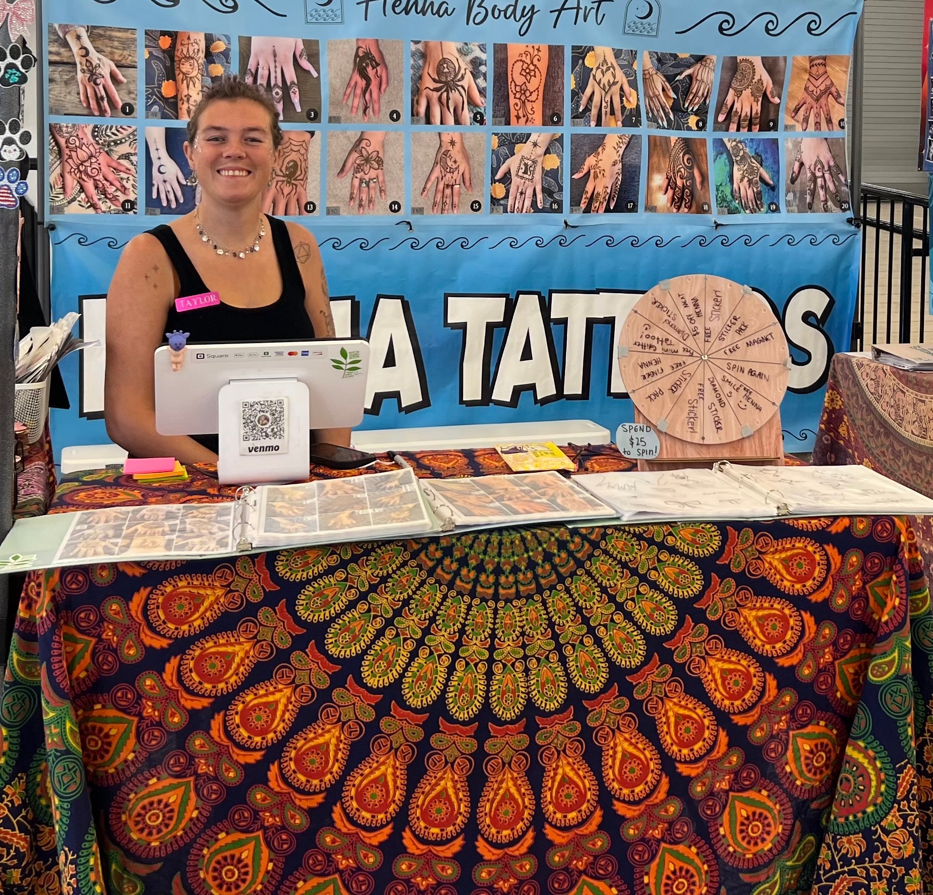 Smiling woman at tattoo booth with colorful fabric tablecloth, display of hand tattoo photos, and a circular artwork.