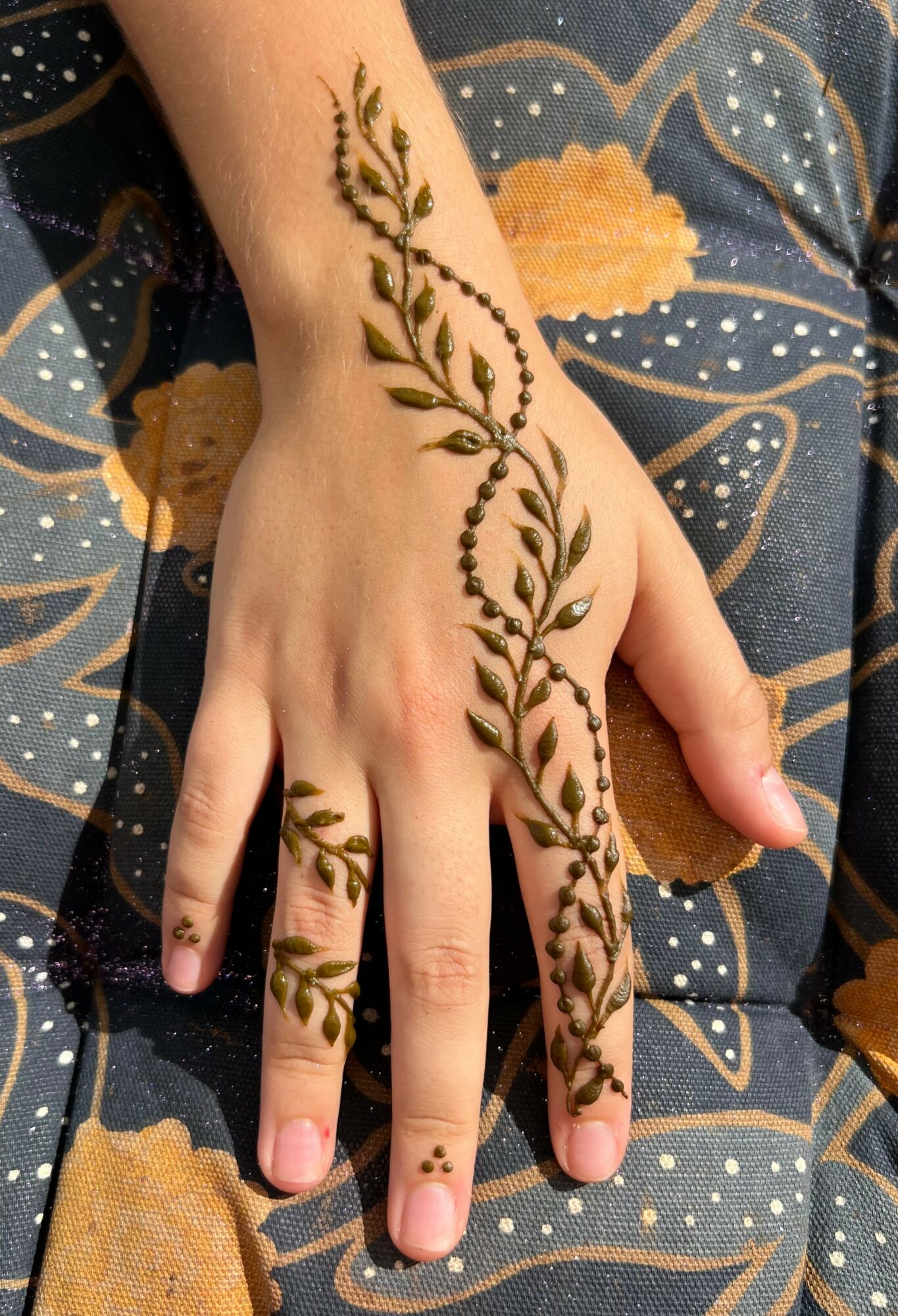 Hand with henna design of leaves and vines on fingers and back of hand, resting on patterned fabric.