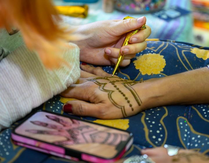Person painting intricate henna designs on another person's hand, with colorful background and a deck of cards nearby.