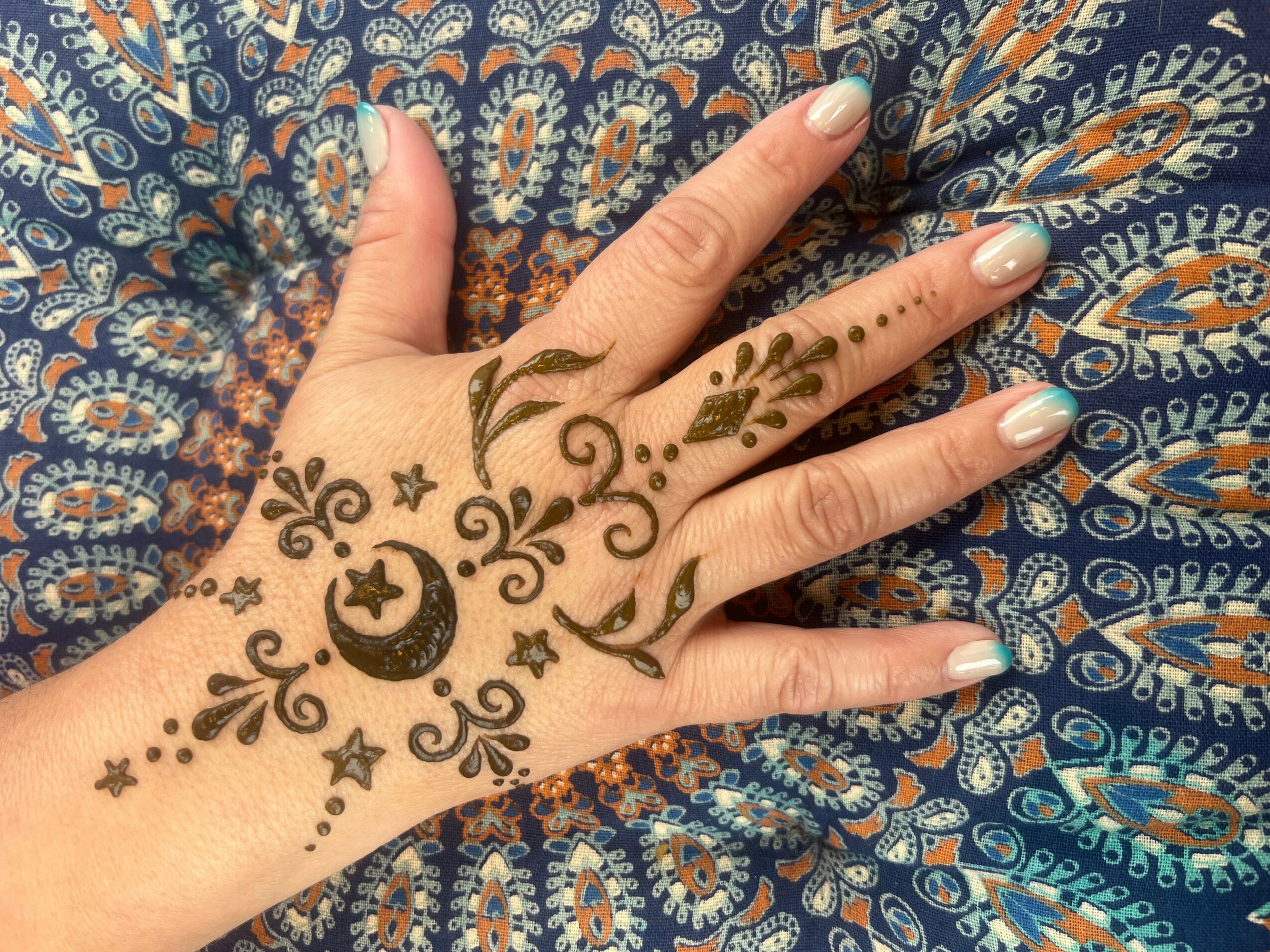 Hand with henna designs on fingers and back, resting on patterned fabric, with a crescent moon, stars, and floral motifs.